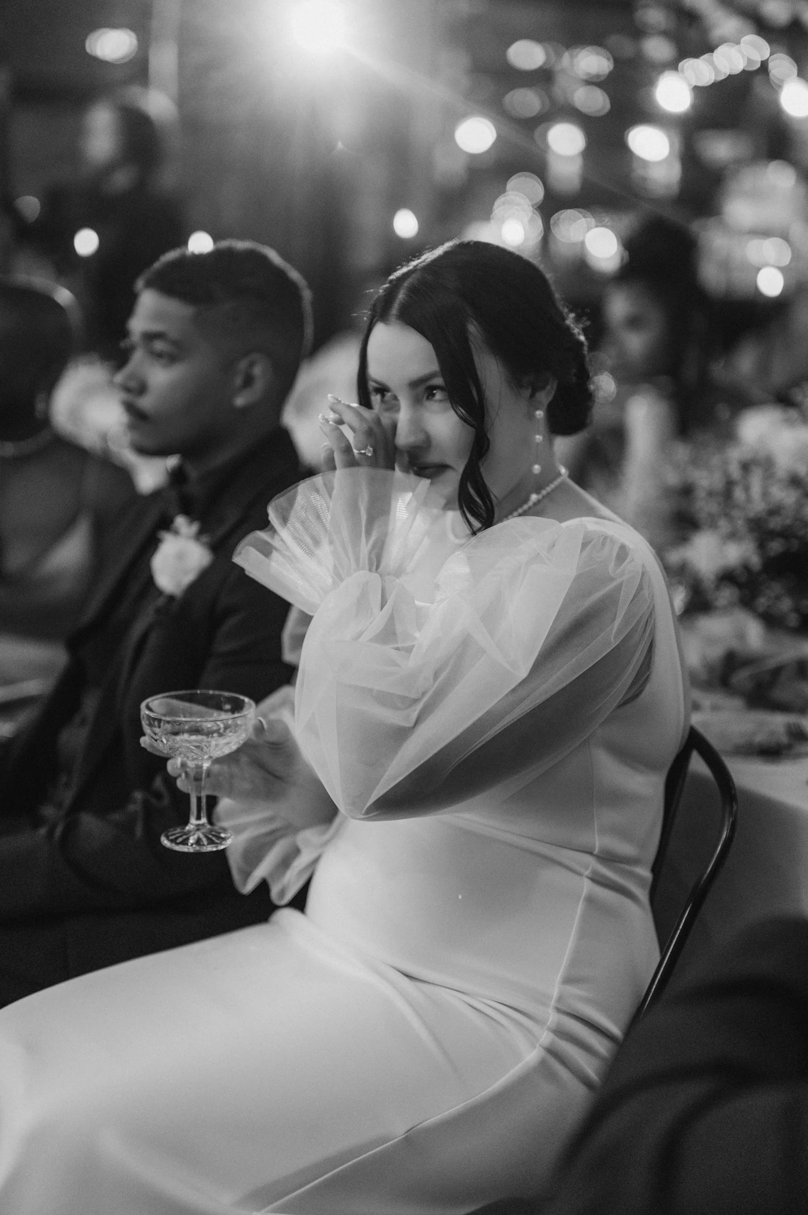 A woman in a white dress, holding a glass and wiping her eye, sitting at a formal event with a man in a dark suit beside her, with a softly lit background.