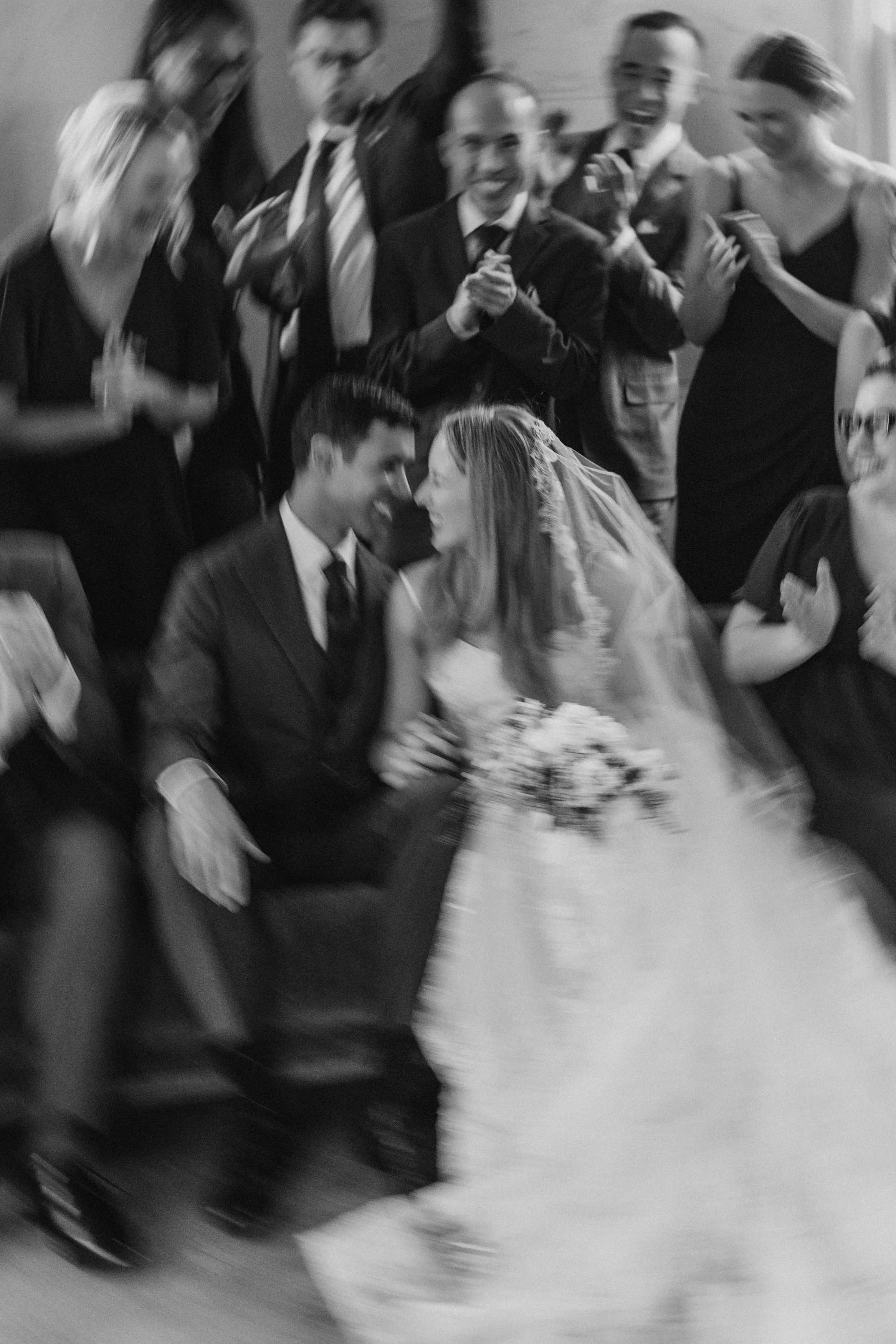 Blurry black and white photo of a wedding scene with a bride and groom sitting close, surrounded by guests celebrating.