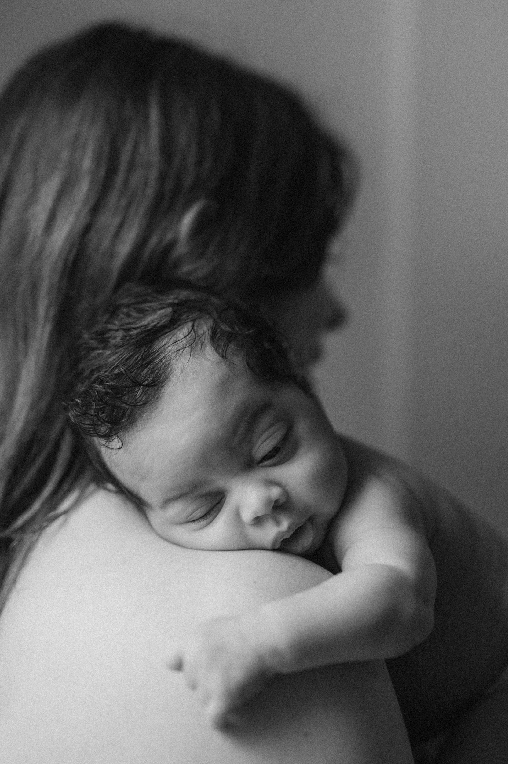 A woman is holding a sleeping baby close to her chest, with the baby's head resting on her shoulder, in a tender moment.