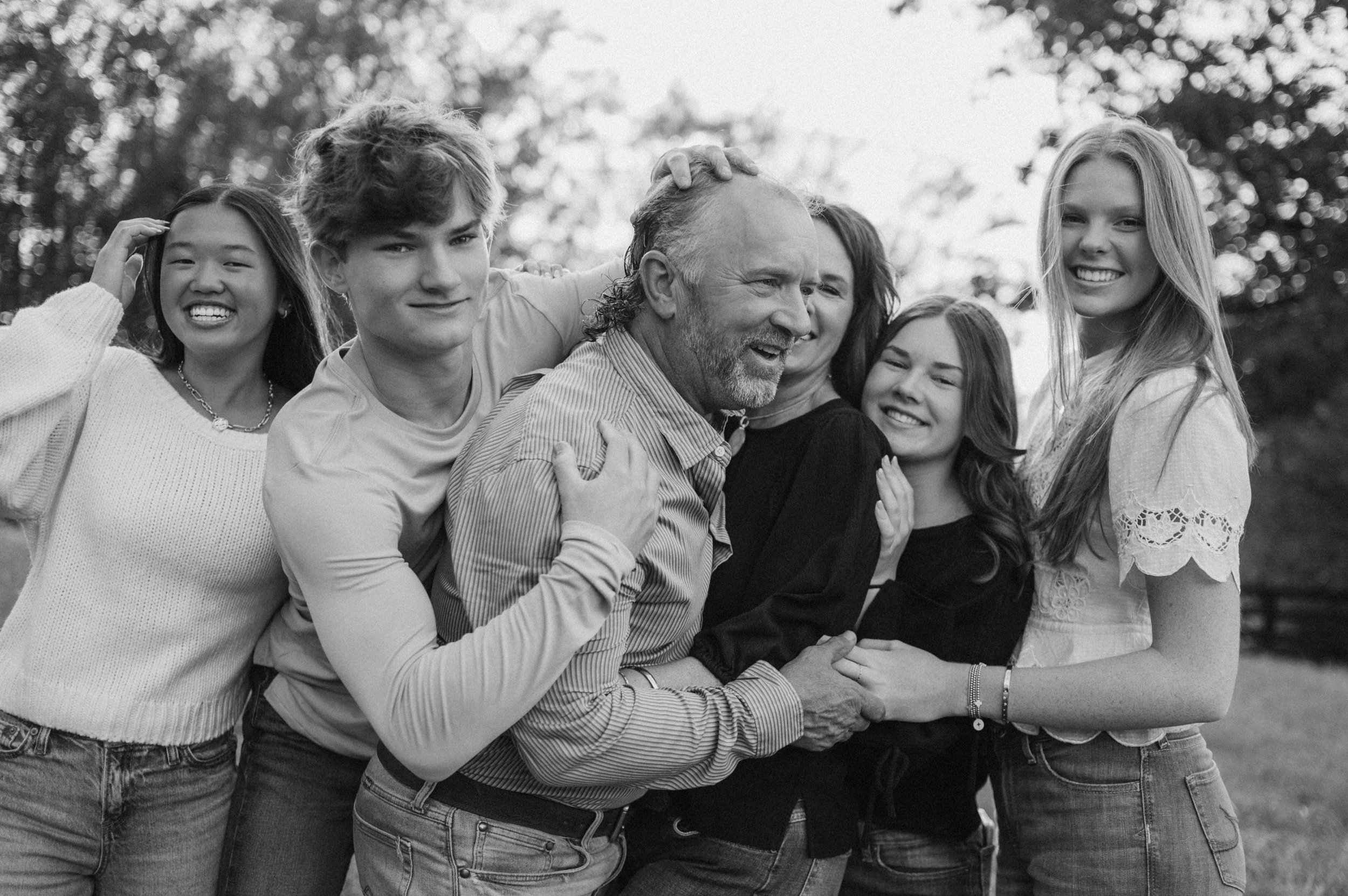 A group of six people, including a man and five women, smiling and embracing outdoors during daytime.