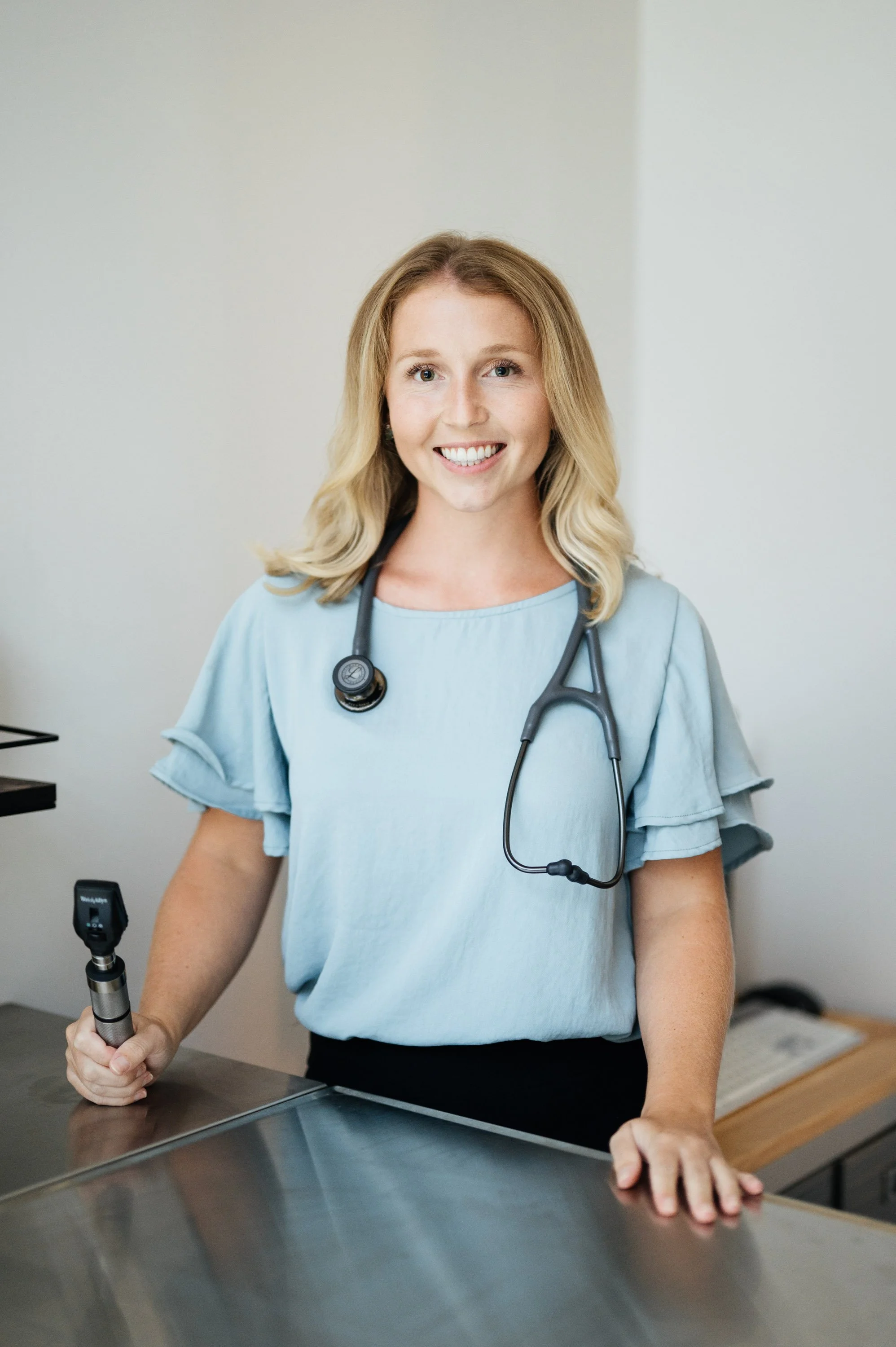 A female healthcare professional wearing a light blue blouse and a stethoscope around her neck, standing at a metal examination table and smiling at the camera.