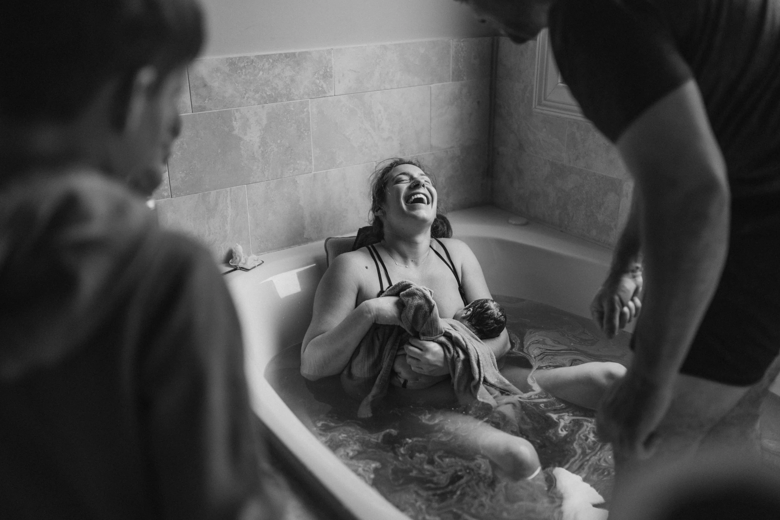 A woman is sitting in a bathtub holding a newborn baby, laughing joyfully. Two people are standing or sitting nearby in a bathroom, observing the scene.