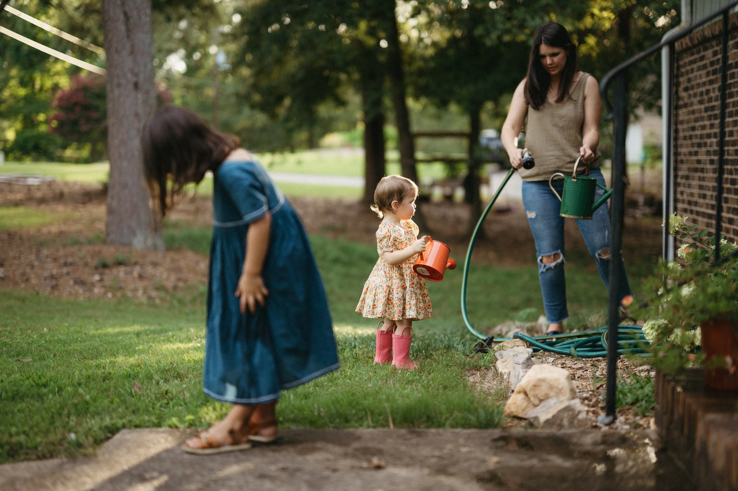 A woman waters plants outside with two young girls, one in a floral dress and pink boots, and the other in a blue dress, on a sunny day in a suburban backyard.