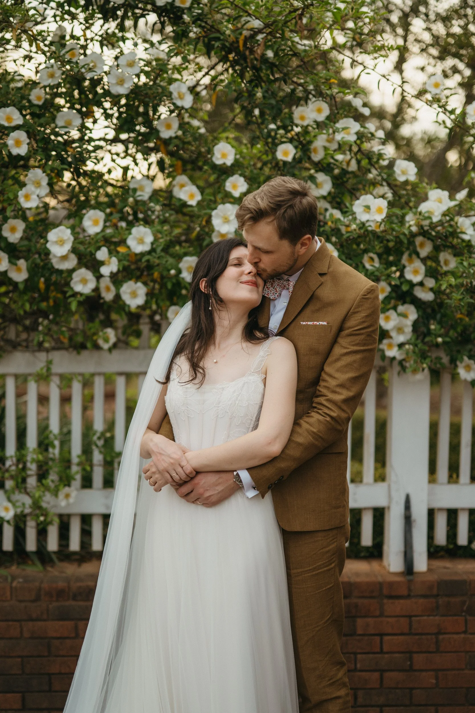 A bride and groom embrace in front of a blooming white flower bush, with a white picket fence and brick wall in the background during their wedding photos.