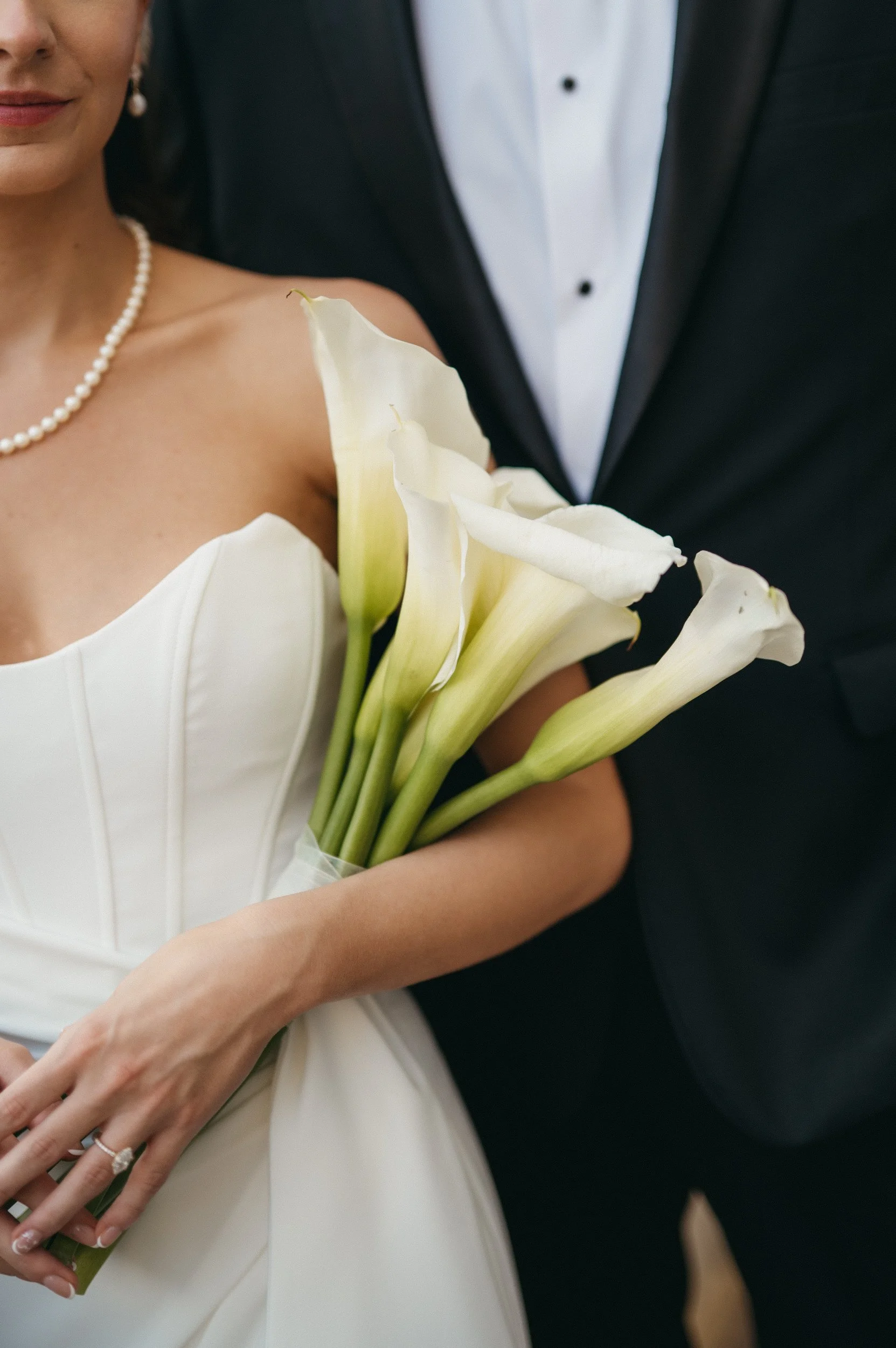 A bride holding a bouquet of white calla lilies, wearing a pearl necklace and a white wedding dress, standing next to a groom dressed in a black tuxedo.