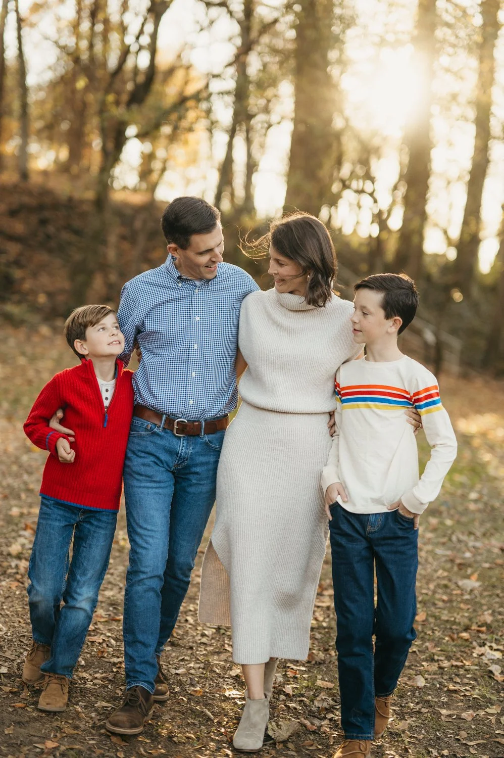 A family of four walking in a wooded park during autumn, with sunlight filtering through the trees. The father and mother are smiling at each other, with the mother wearing a white sweater dress. The two sons, one in a red sweater and jeans, and the 