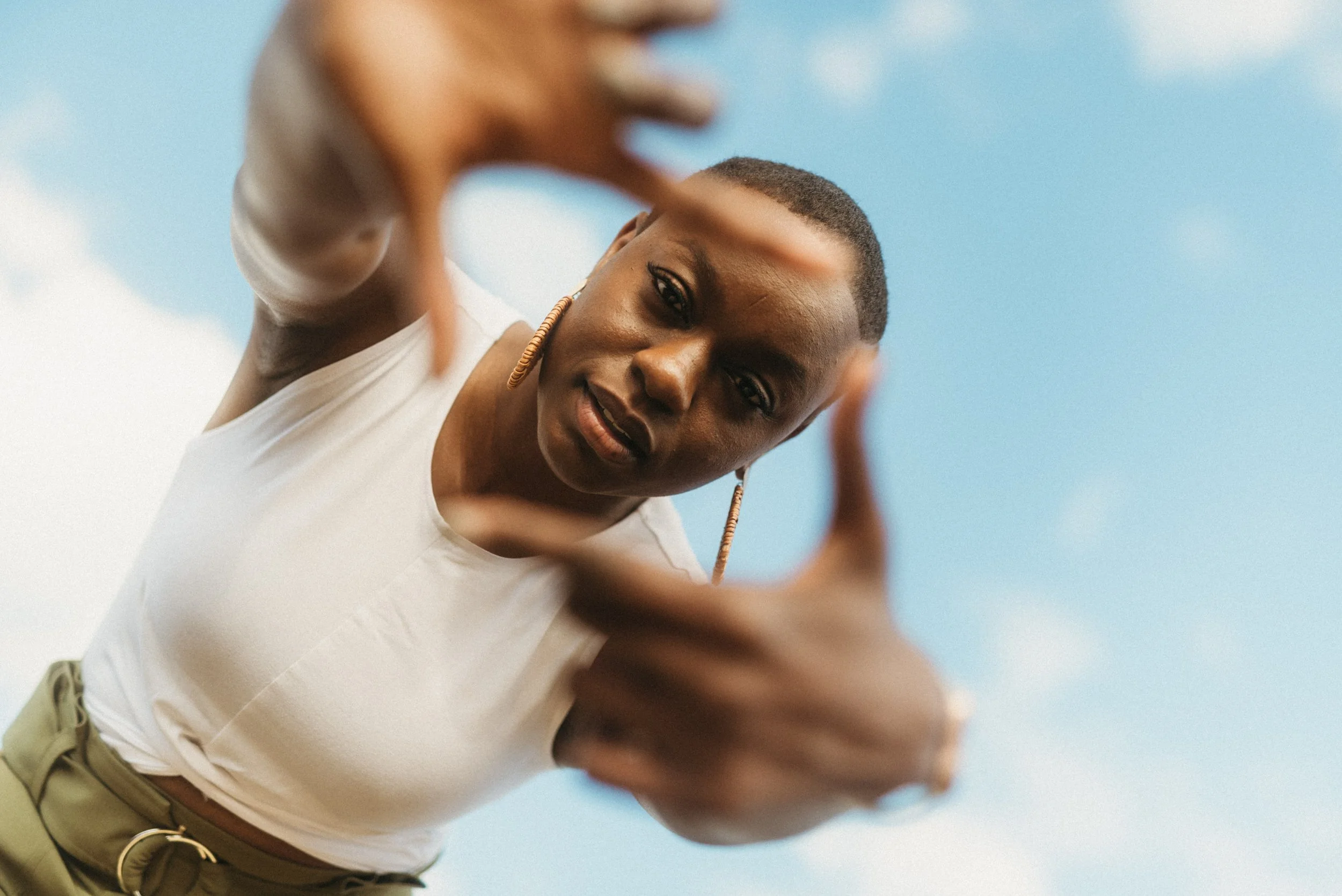 A woman with a short hairstyle and hoop earrings looks into the camera through a heart-shaped frame formed with her hands, against a bright blue sky with some clouds.