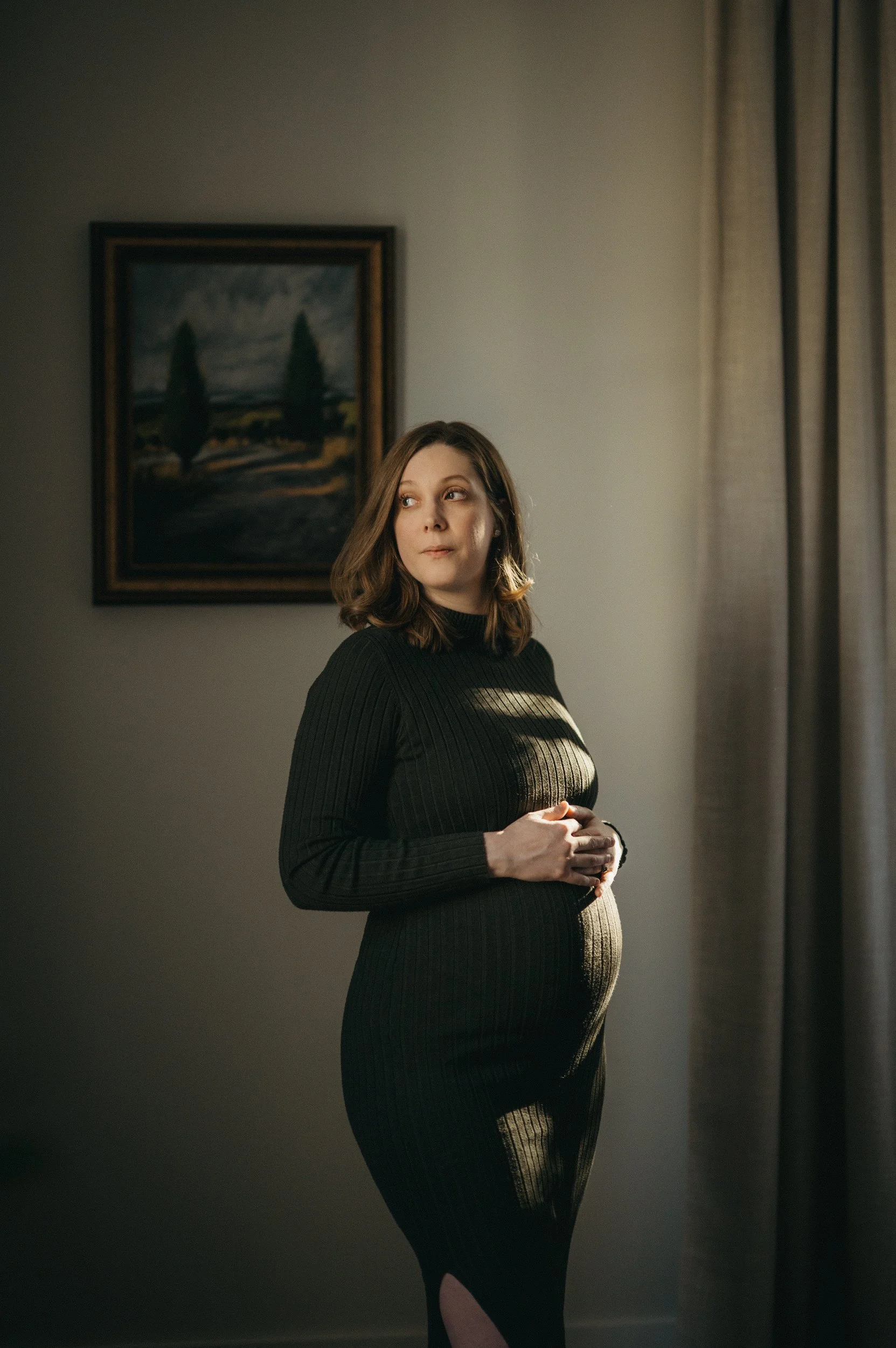 A pregnant woman in a black ribbed dress standing near a window with curtains, holding her belly and looking to the side.