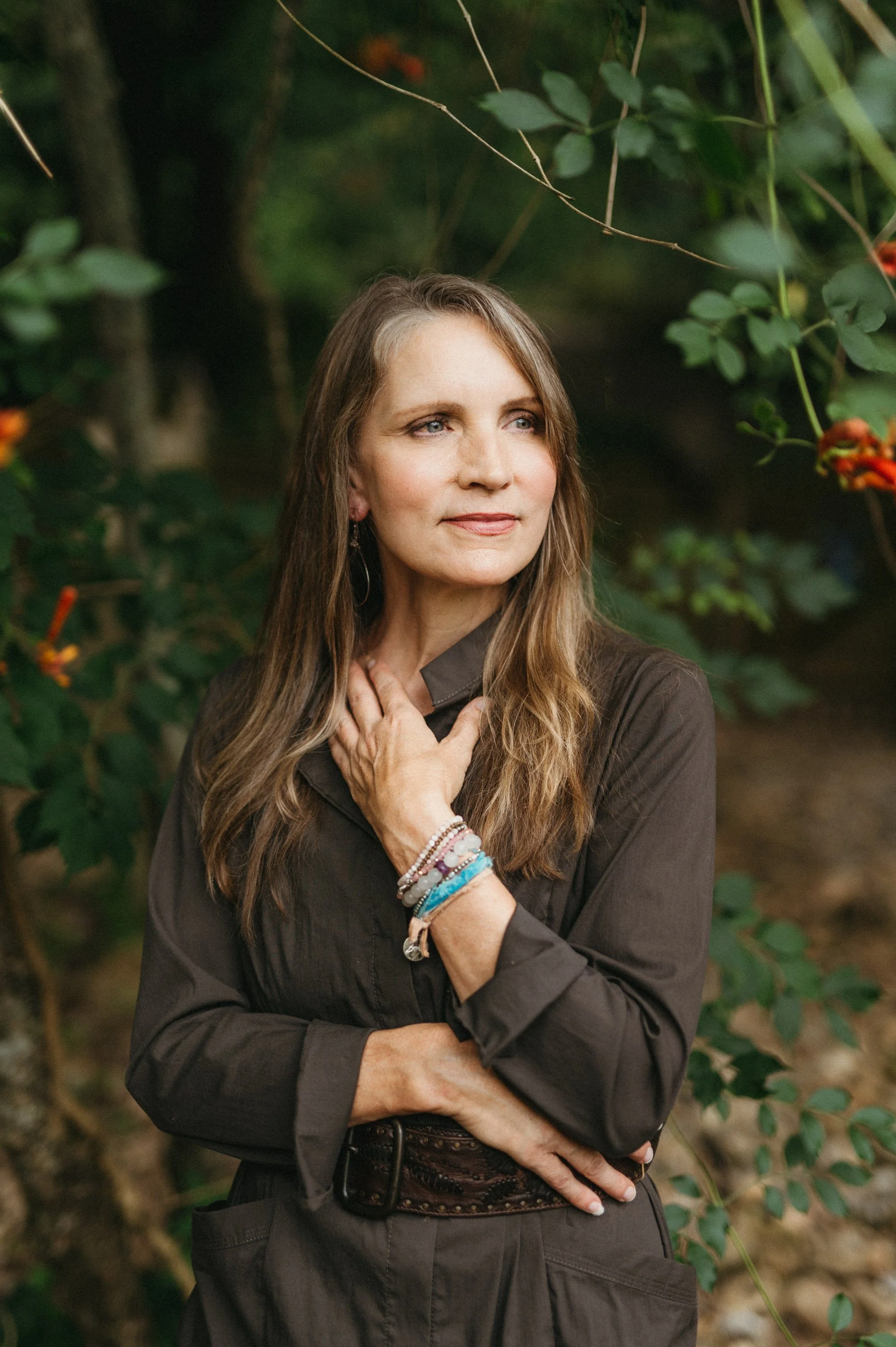 A woman with long brown hair, wearing a dark brown jacket and bracelets, standing outdoors among green leaves and branches, looking off to the side.