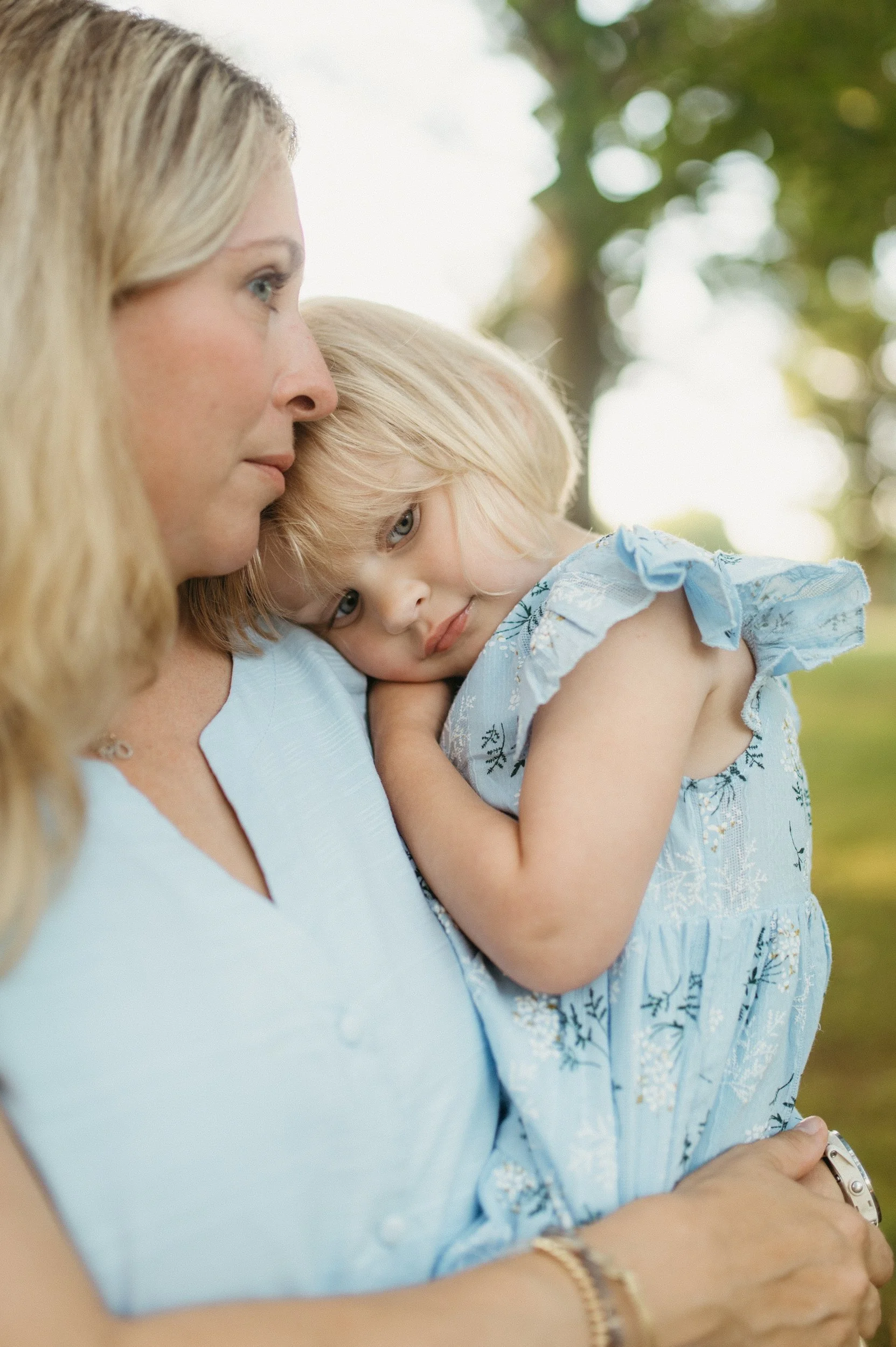 A woman holding a young girl close with the girl resting her head on the woman's chest, outdoors with blurred trees in background.