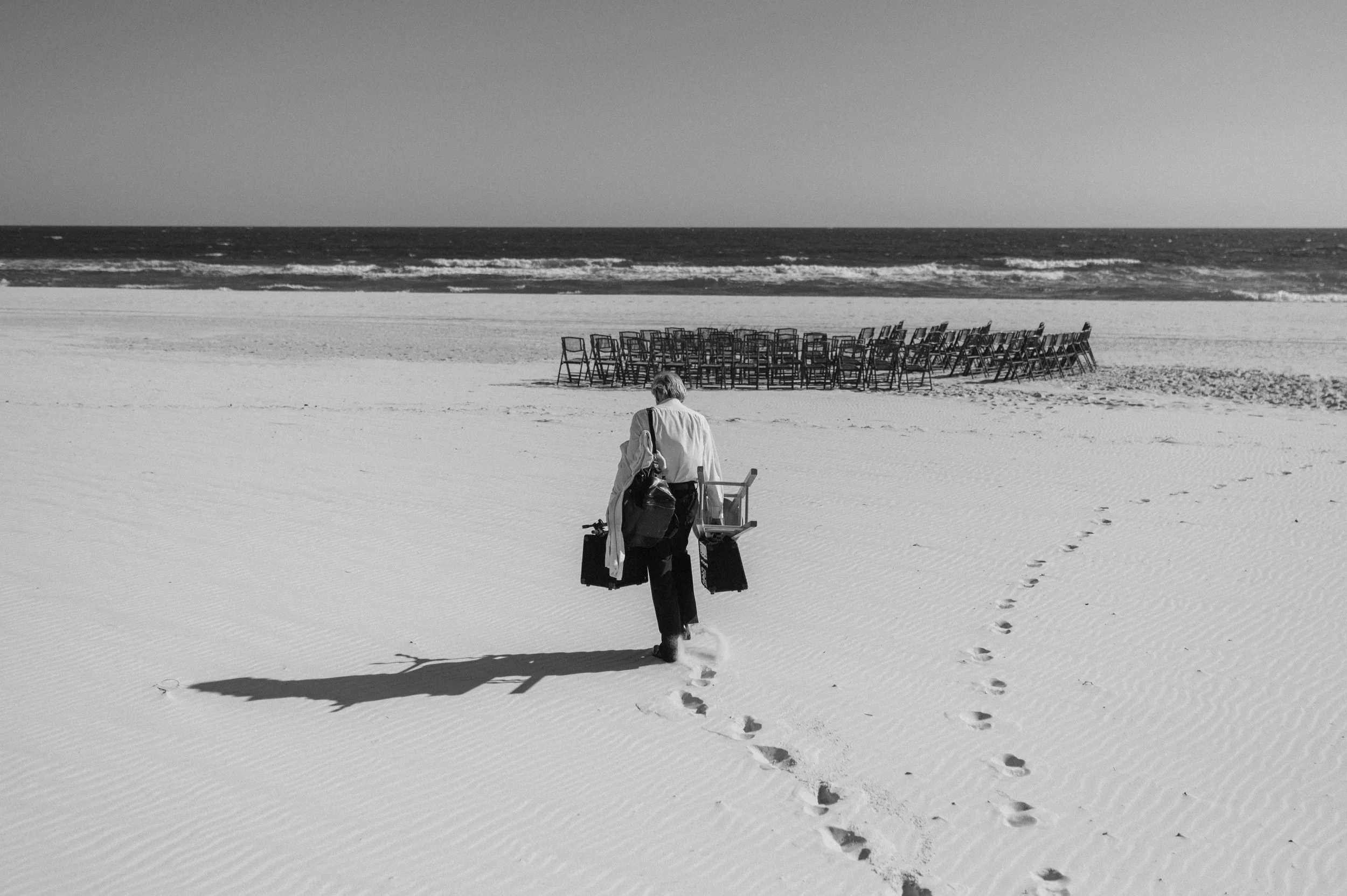 A person walking on a sandy beach carrying luggage and chairs, with numerous chairs stacked in the background near the shoreline.