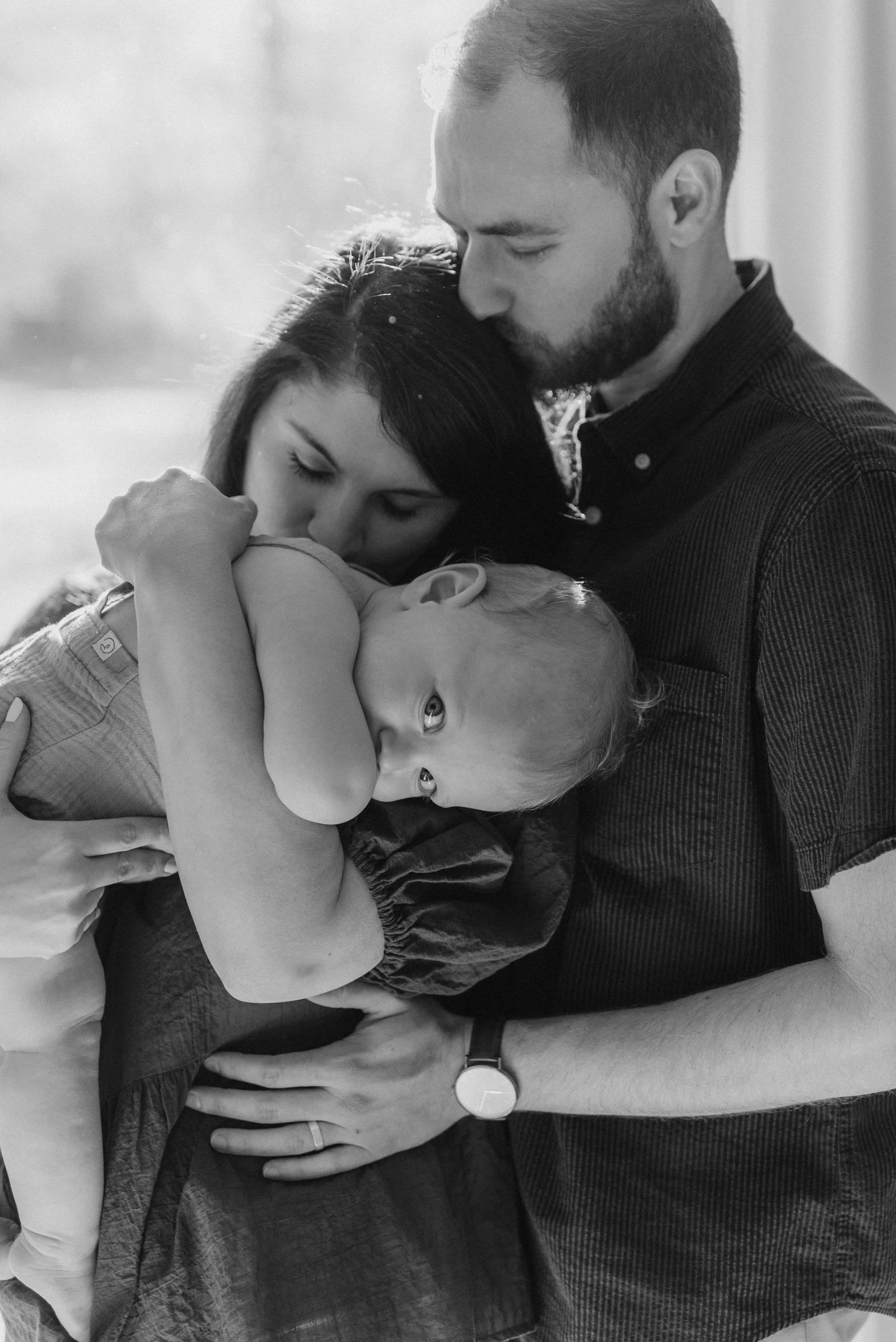 A black and white photo of a family moment with a man, woman, and young child. The woman is holding the child close, while the man leans in for a gentle kiss on the woman's head. The child is resting peacefully on the woman's chest, looking at the camera with a curious expression.