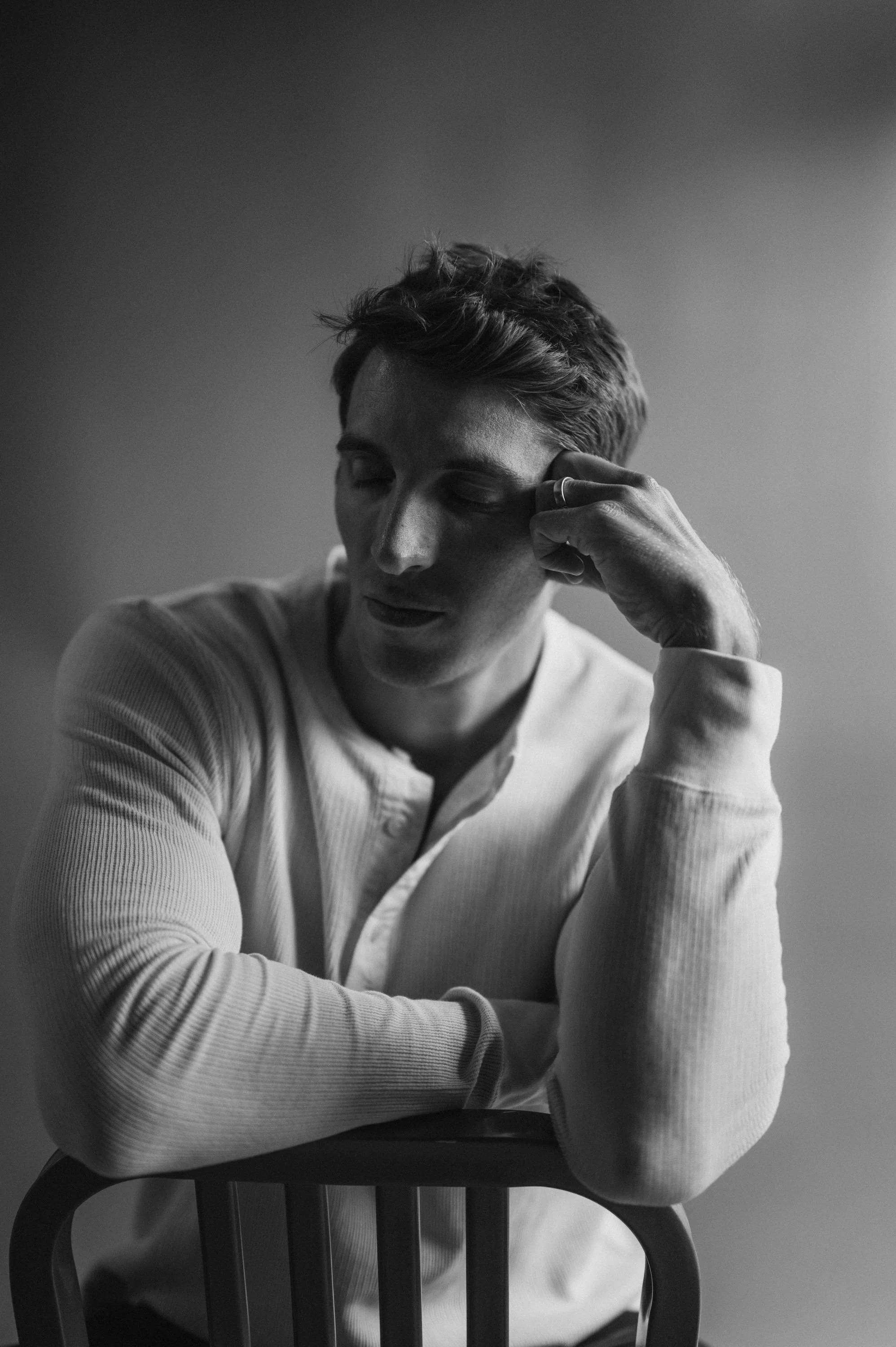 A black and white portrait of a young man with short hair, sitting on a chair with arms crossed on a table. He is wearing a long-sleeve shirt and is resting his head on his hand with eyes closed, appearing thoughtful or relaxed.