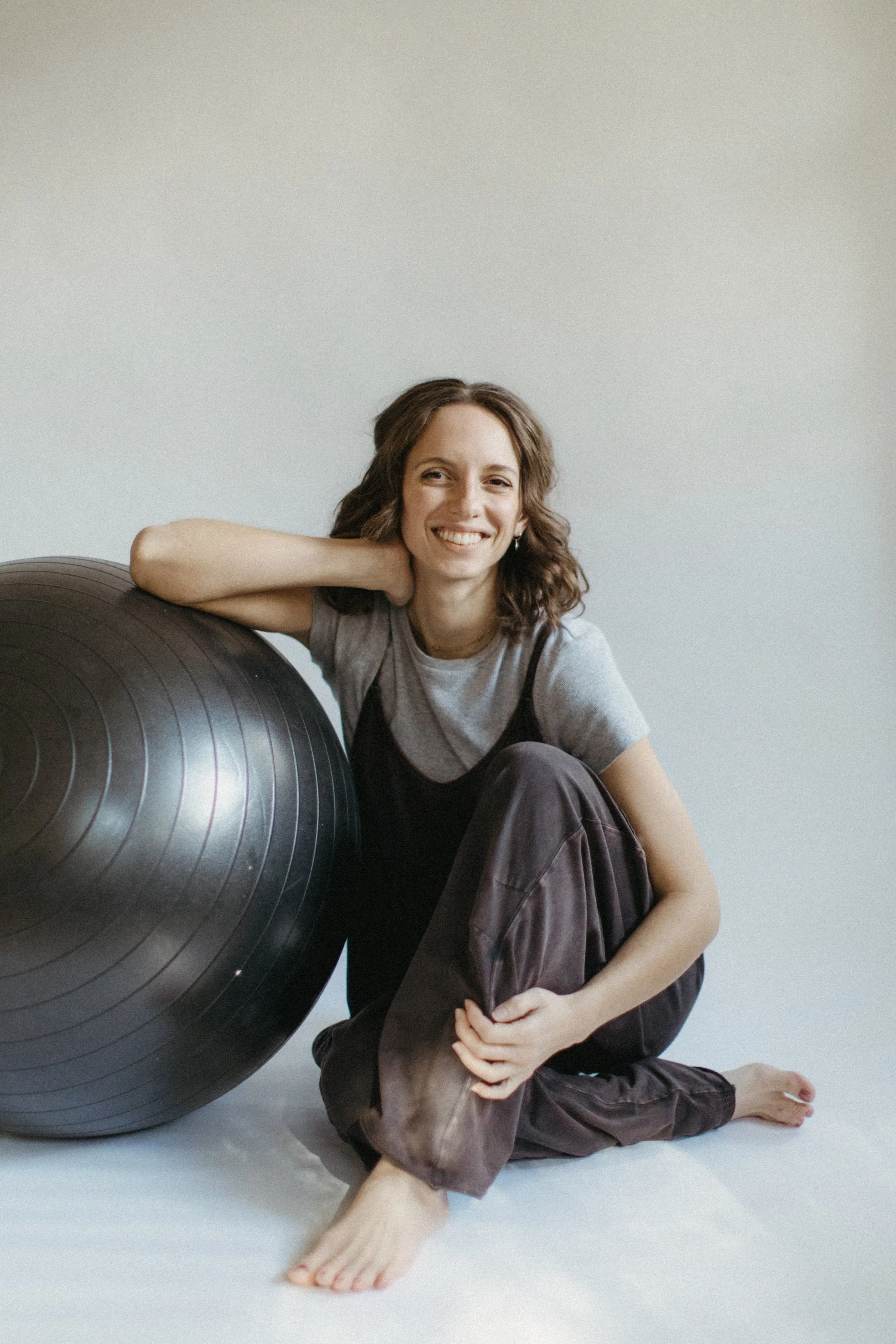 A woman sitting on the floor next to a large exercise ball, smiling and resting her head on her arm.