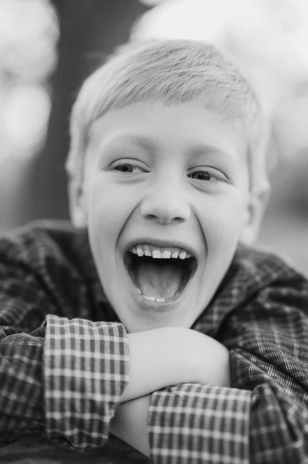 Black and white photo of a young boy smiling broadly with mouth open, wearing a plaid shirt, arms crossed in front of him.