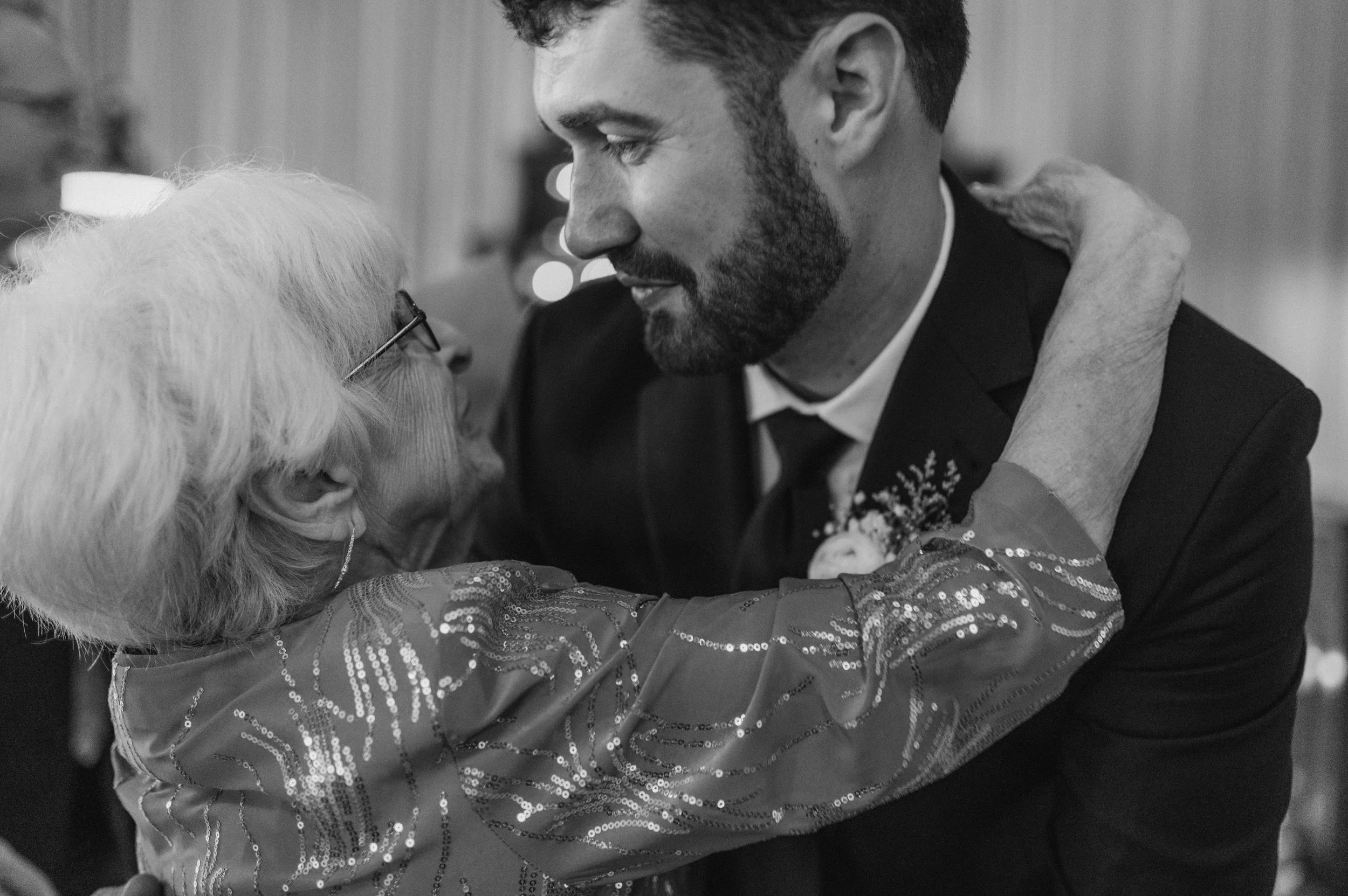 An elderly woman with white hair and glasses embraces a young man in a suit during a dance or celebration, both appearing happy and close.