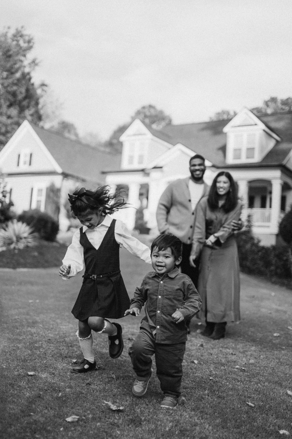 Two young children, a girl and a boy, joyfully running and playing on a grassy front yard of a house, with a smiling couple standing behind them. The scene is in black and white.
