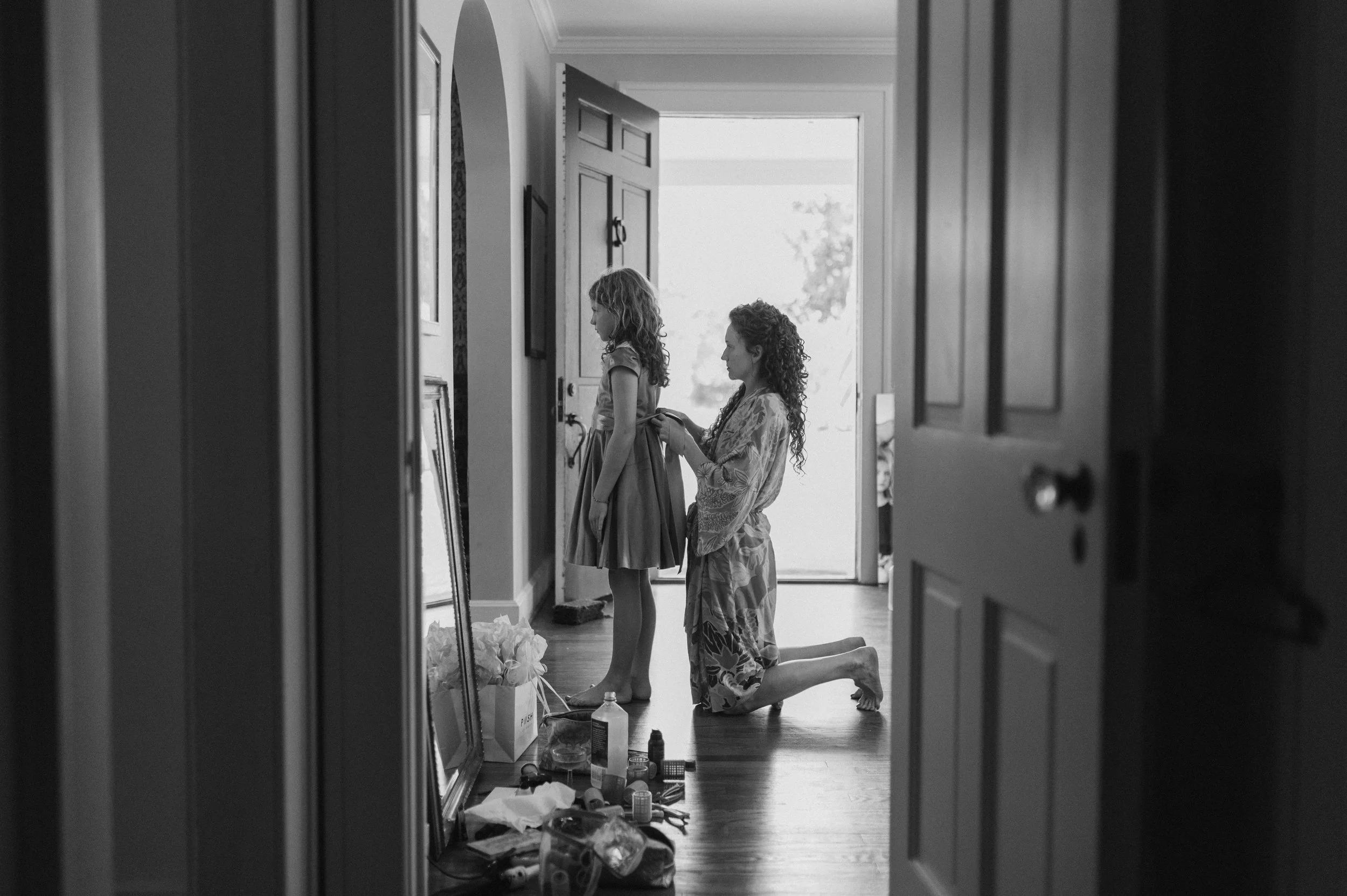 A woman kneeling on the floor helping a young girl prepare for an event in a hallway, with the front door open in the background. Items and a framed mirror are scattered on the floor.