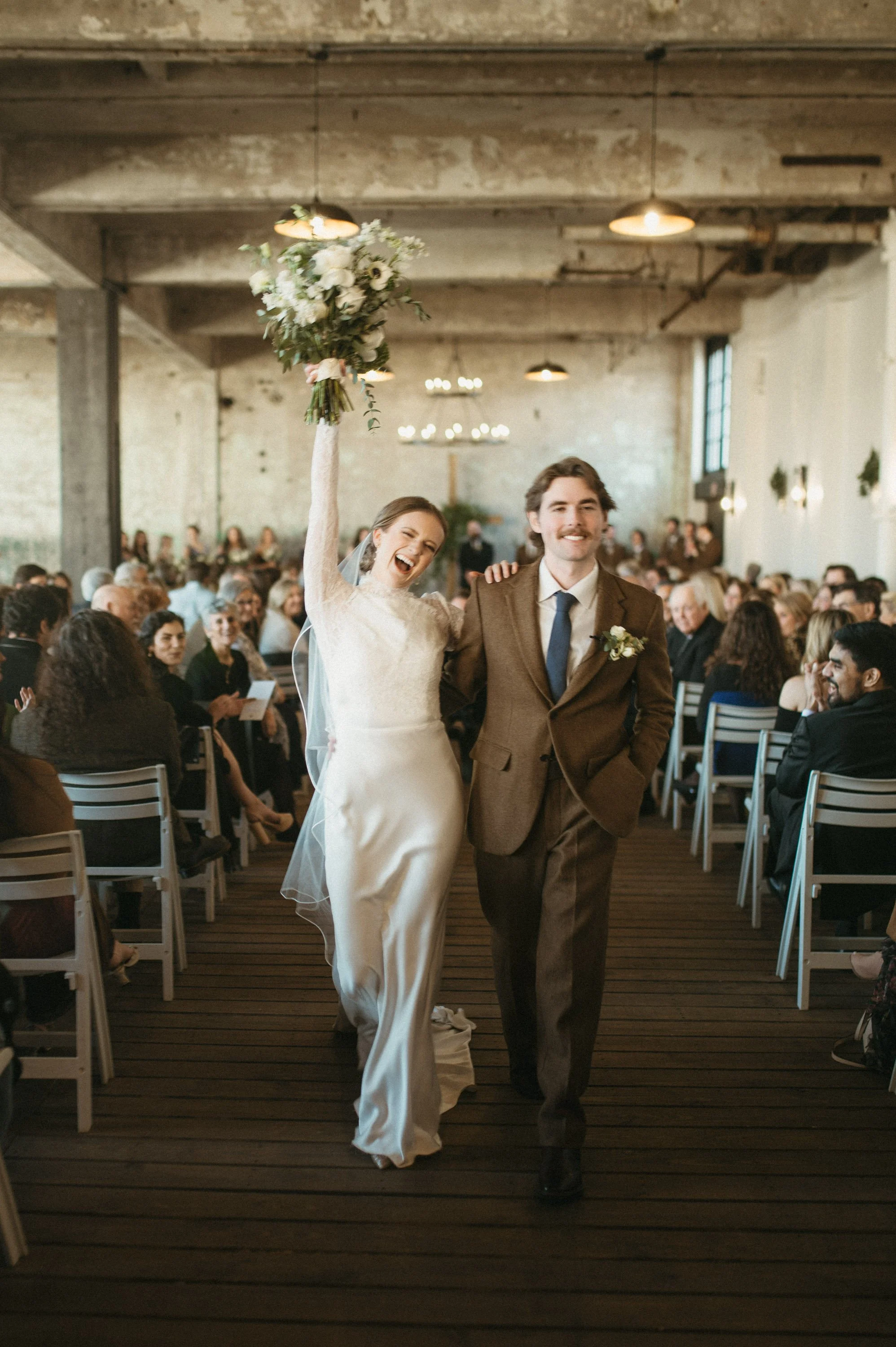 A newlywed couple walking down the aisle at their wedding ceremony, the bride holding a bouquet of flowers raised in the air, with guests seated and smiling in the background.