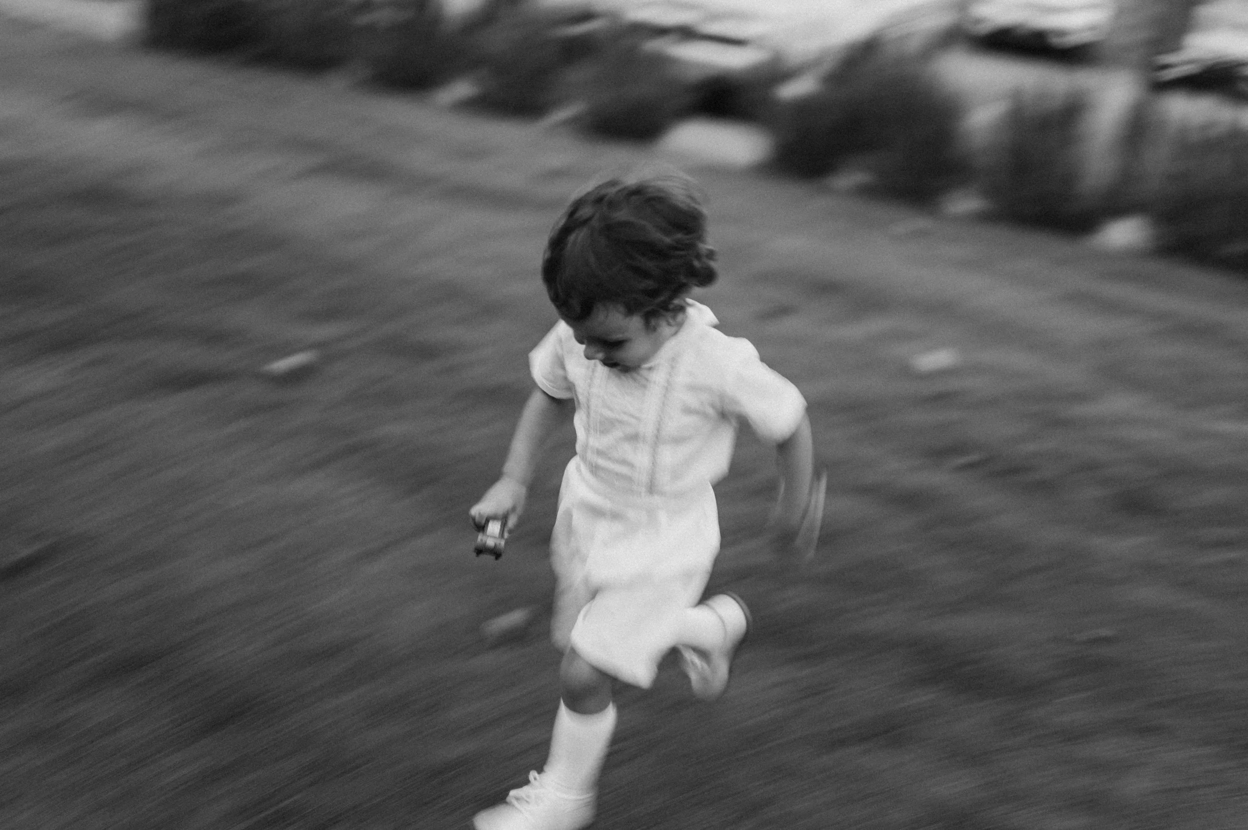 Child in a white dress running with a small toy car in hand on a blurred grassy field in black and white.