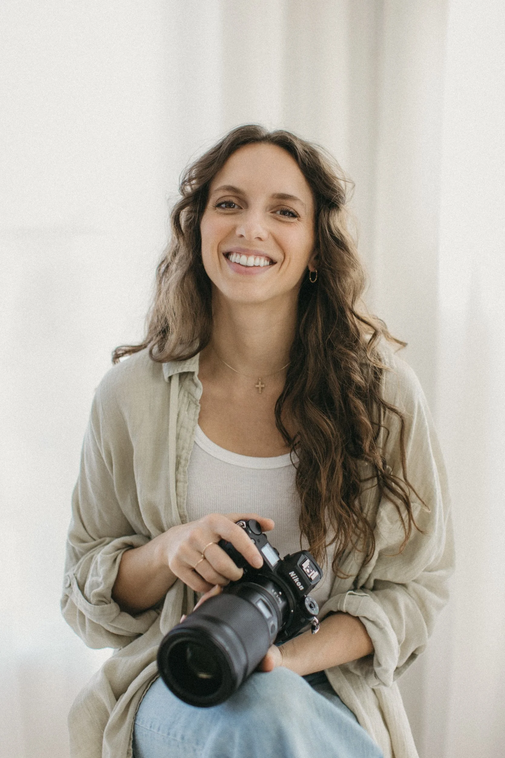 A young woman with long, wavy brown hair sitting and holding a Nikon camera, smiling at the camera, indoors with light-colored background.