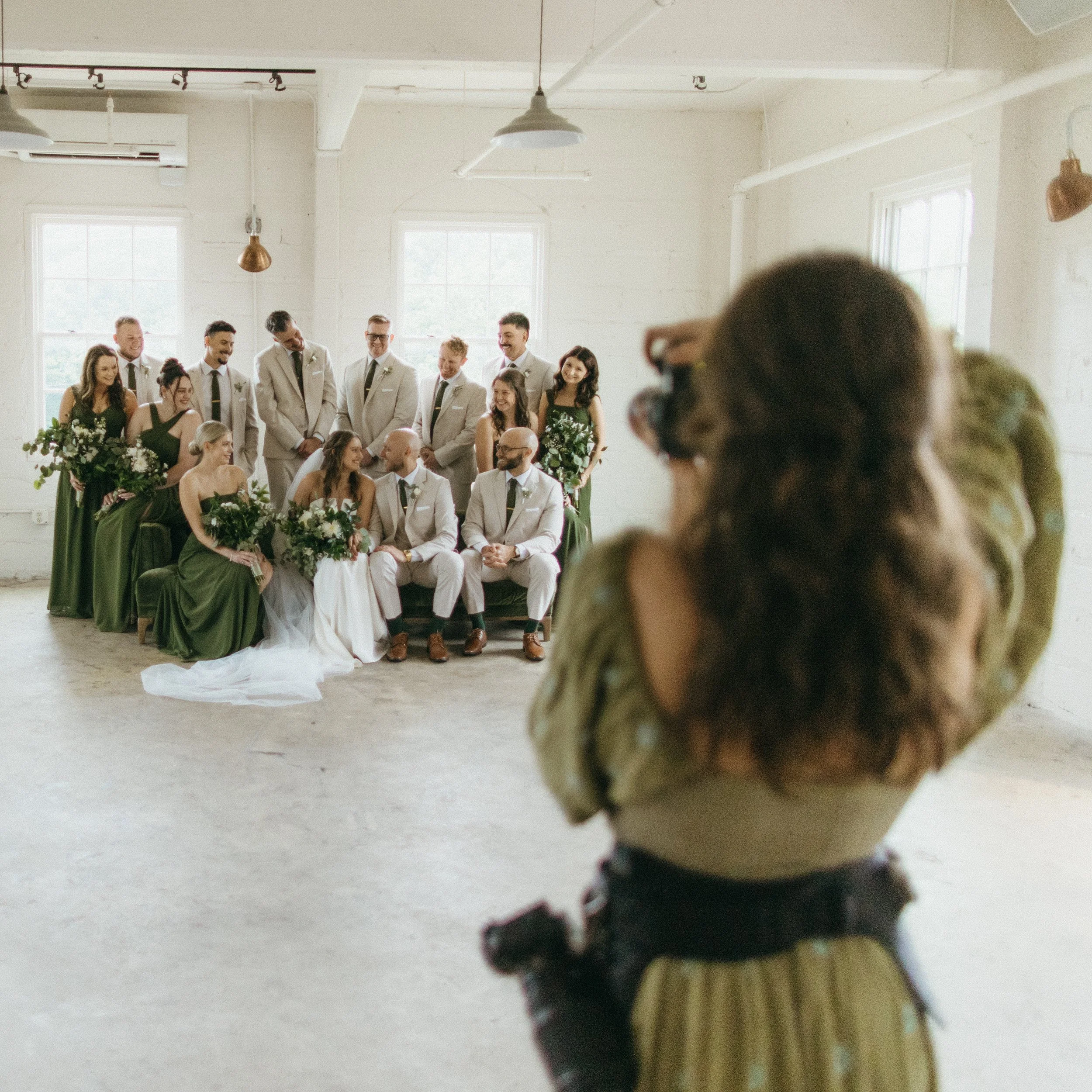 A wedding group photo with the bride, groom, bridesmaids, and groomsmen in a bright, industrial-style room. The bride and groom sit in the front with bridesmaids and groomsmen standing behind them, all dressed in formal attire.