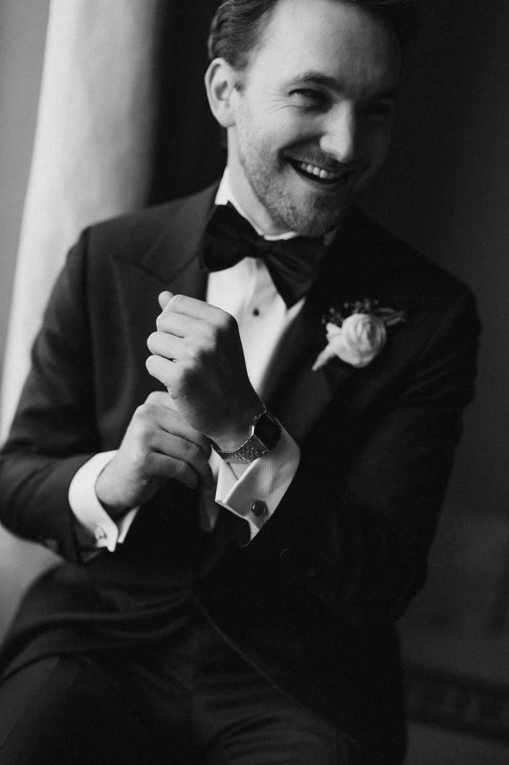 A man in a tuxedo and bow tie smiling joyfully, with a boutonnière on his lapel, adjusting his cufflink.