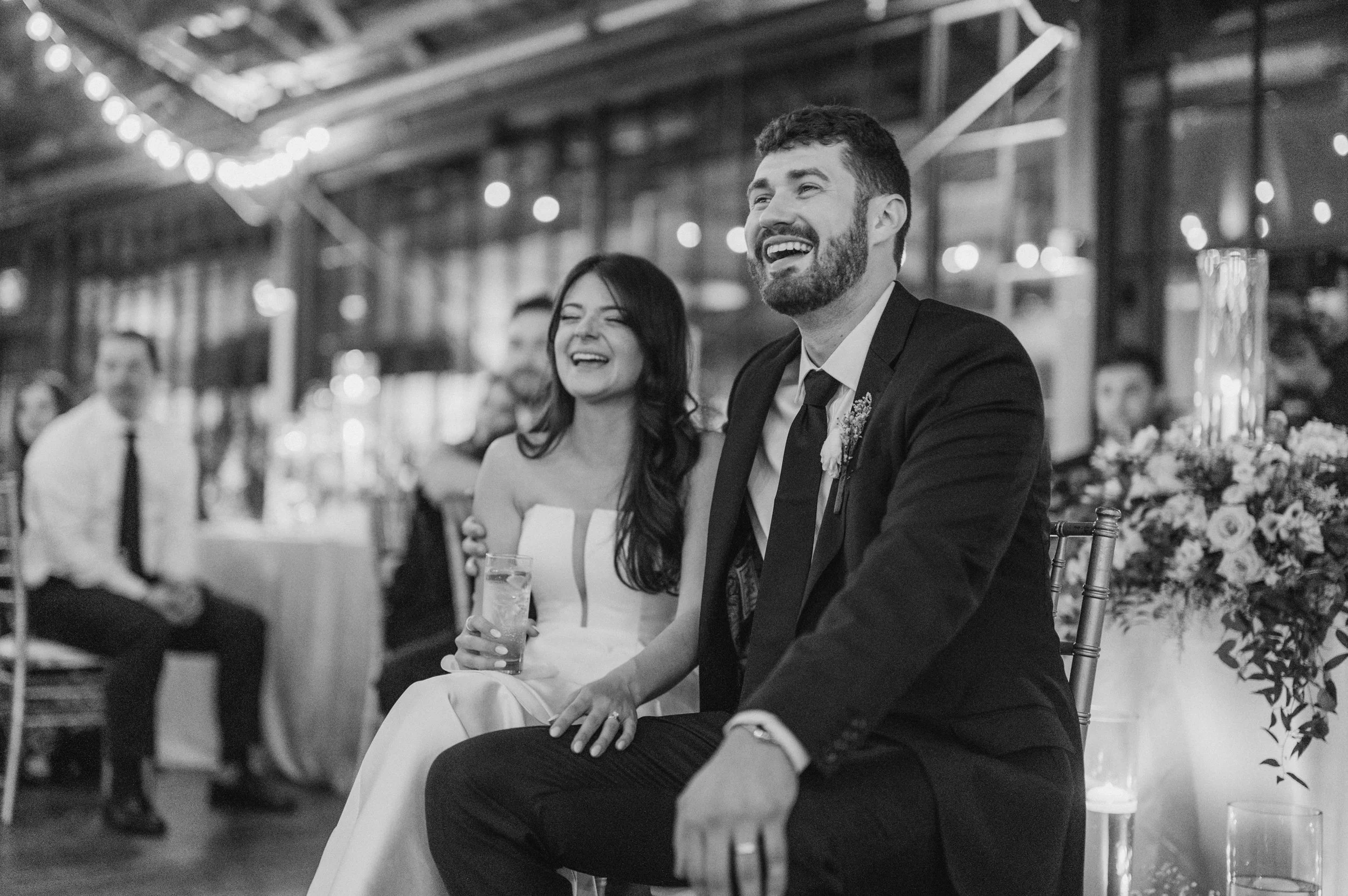 A black-and-white photo of a smiling bride and groom sitting together during their wedding reception, with guests in the background.