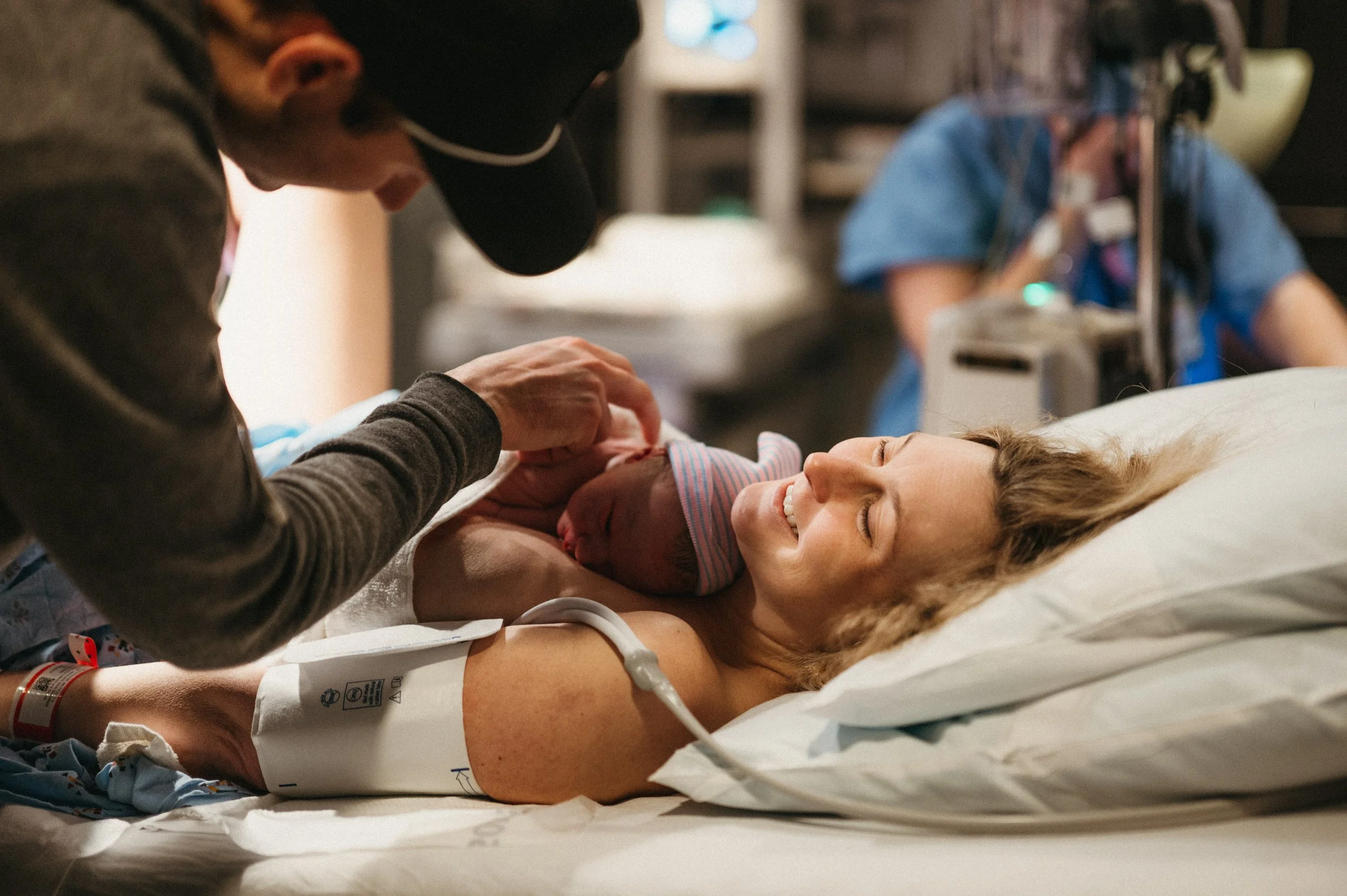 A woman lying in a hospital bed, holding a newborn baby, with a person in a cap leaning over to gently touch the baby's hand, in a hospital room.