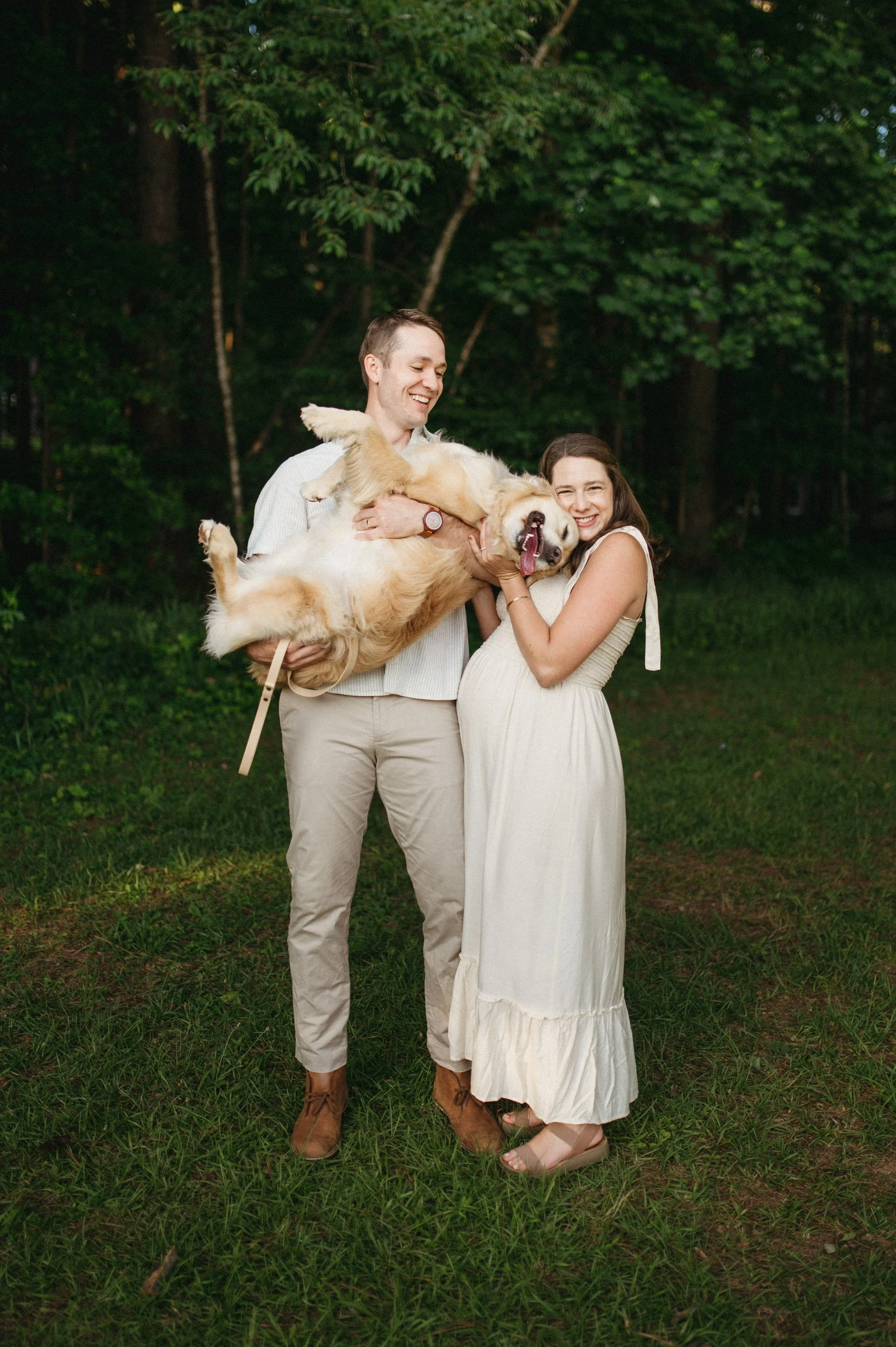 A happy couple with a dog outdoors in a wooded area, the man holding the dog and the woman hugging it, all smiling.