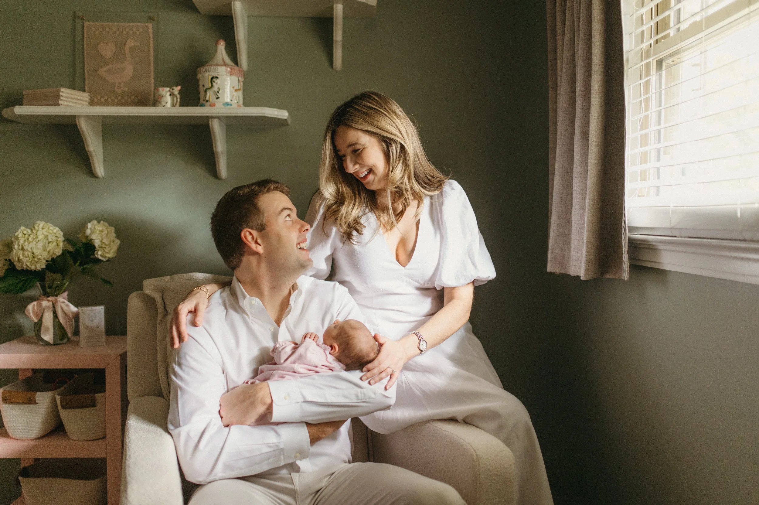 A happy family of three with a newborn baby in a cozy living room, smiling and looking at each other.