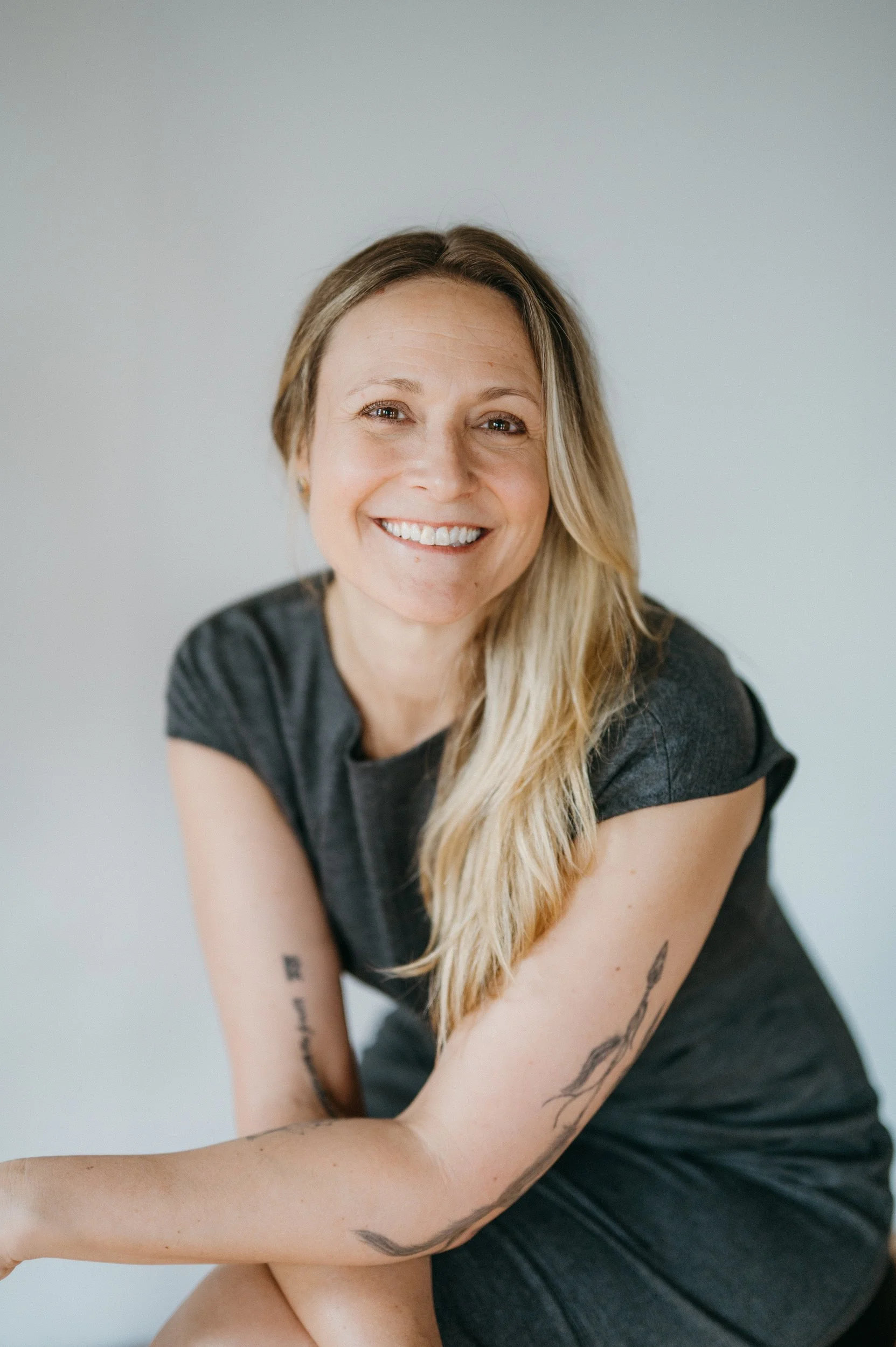 A smiling woman with long blonde hair wearing a dark gray t-shirt, sitting against a plain light-colored background.