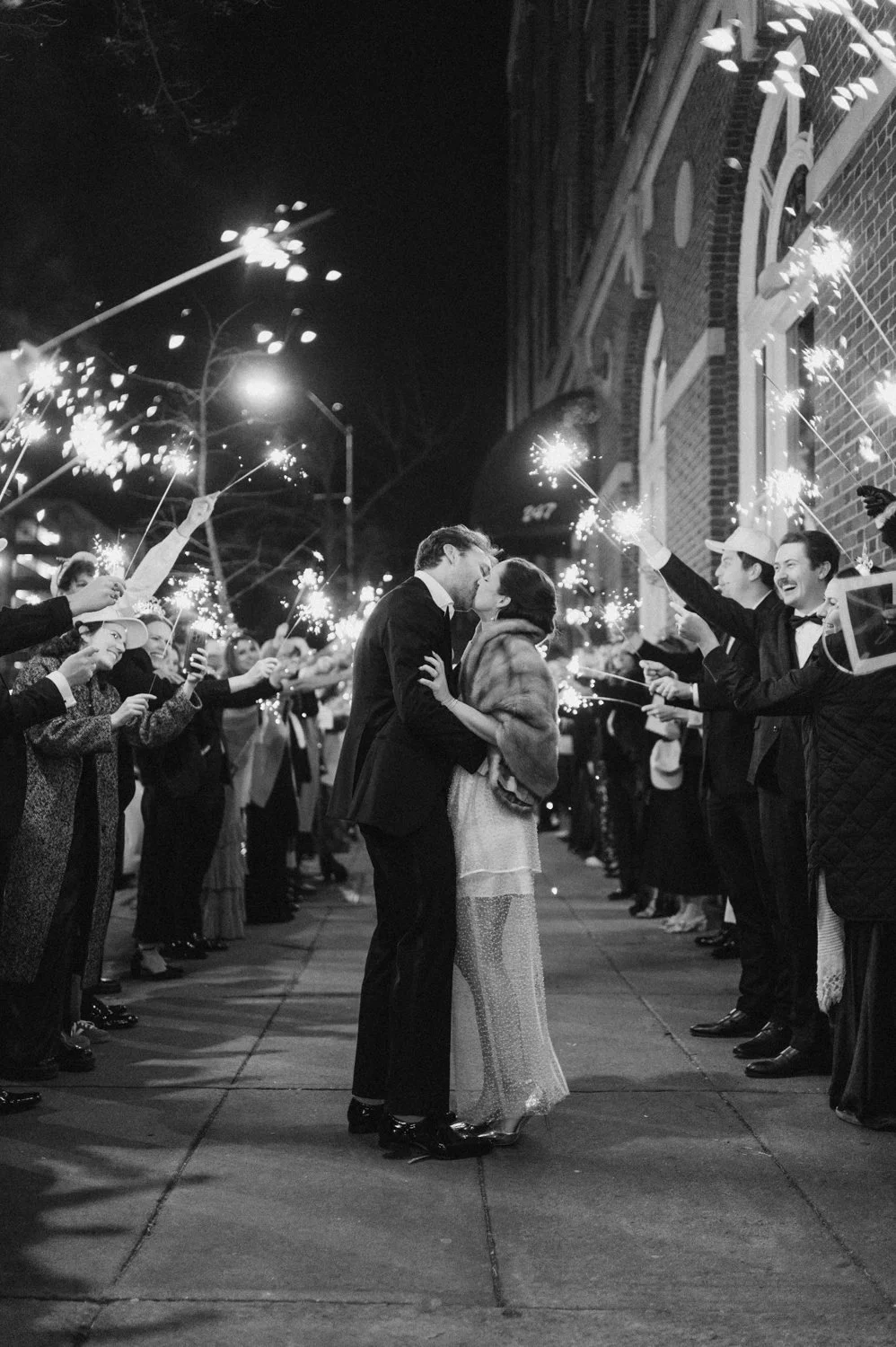 A couple is kissing in the middle of a street at night surrounded by people holding sparklers, celebrating a joyful event, possibly a wedding.