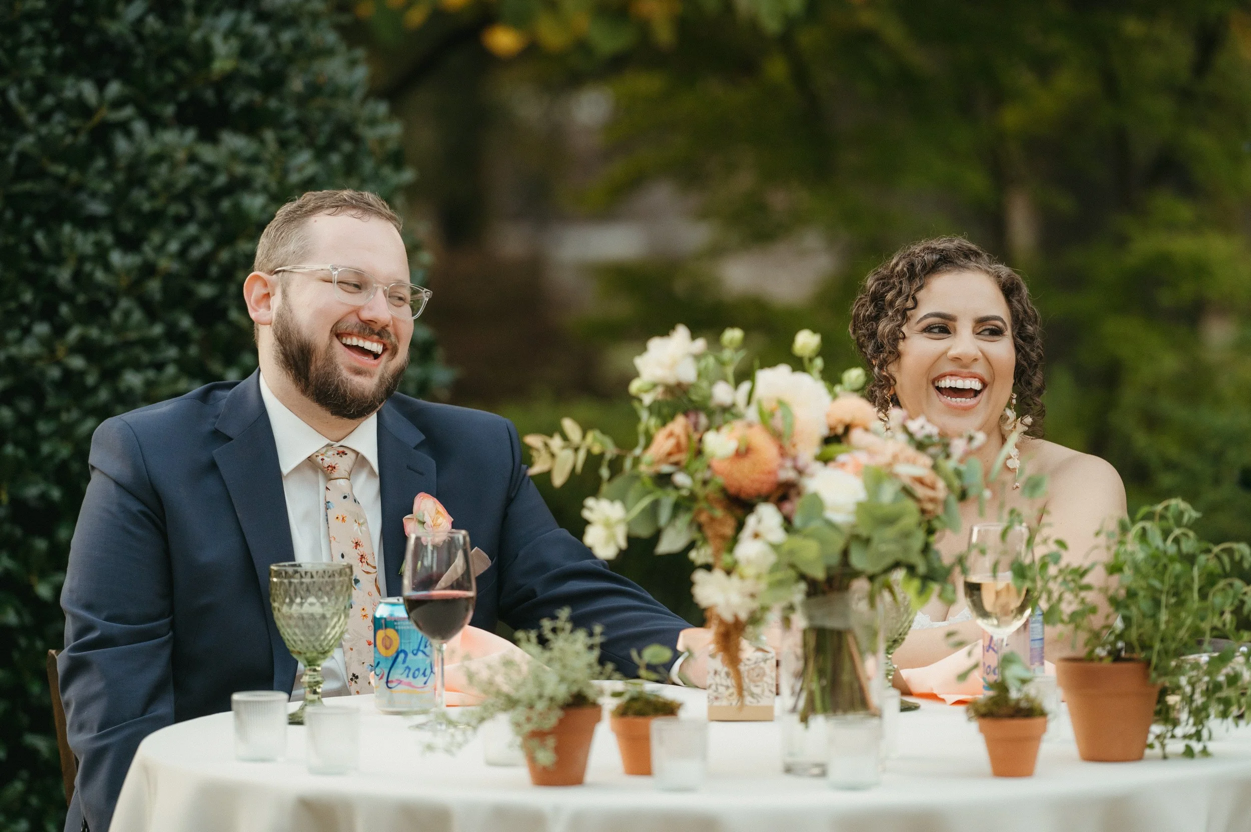 A smiling couple at a wedding reception, sitting at a table with a large floral centerpiece, glasses of wine, and potted plants, outdoors with green trees in the background.