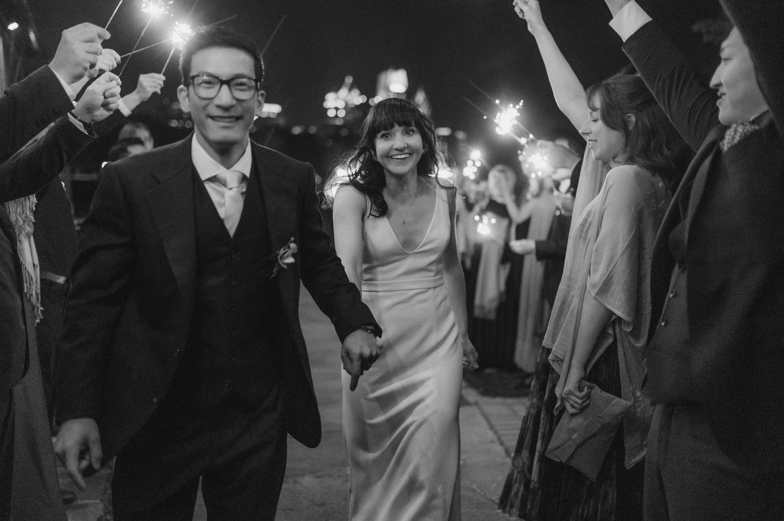 A couple dressed in formal attire holding hands and smiling during a fireworks display at night with friends in the background.