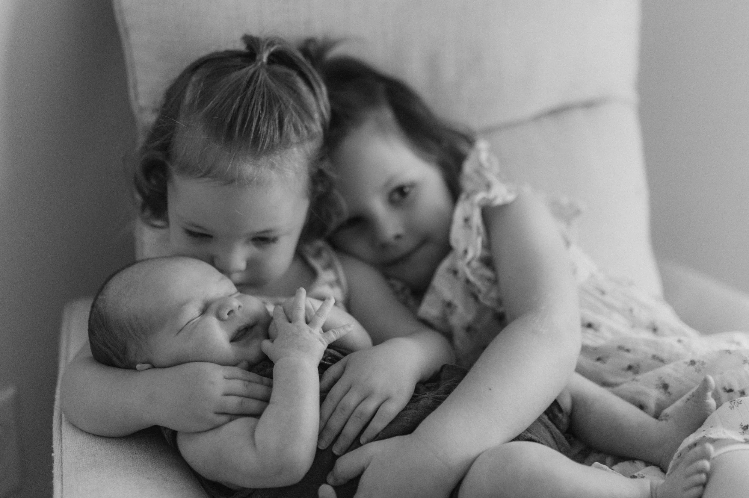Two young girls and a baby lying on a couch, with the baby sleeping and the older children looking at the baby.