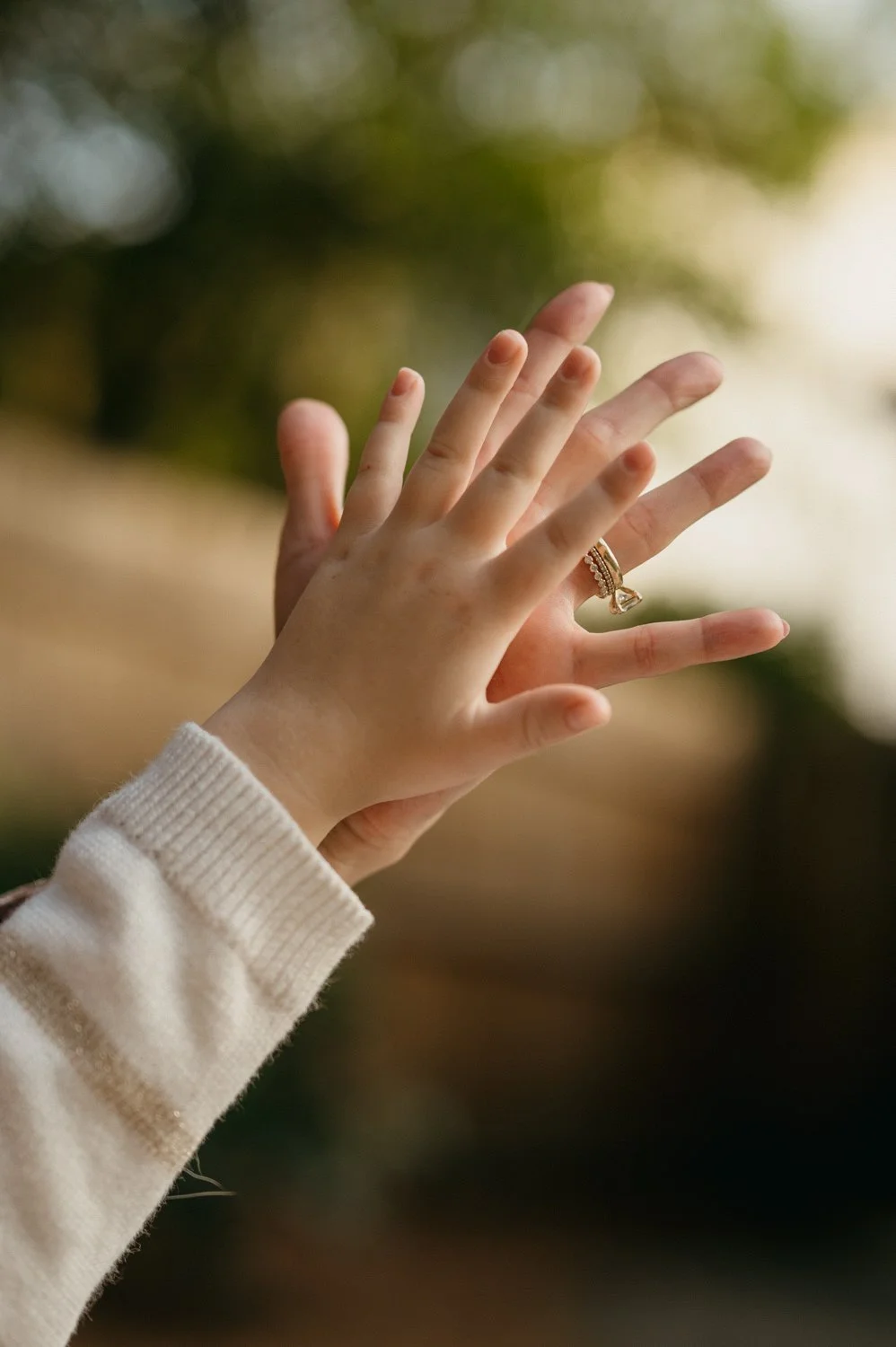 Close-up of two hands, one adult and one child, with the adult wearing a gold ring, reaching towards a blurred outdoor background.