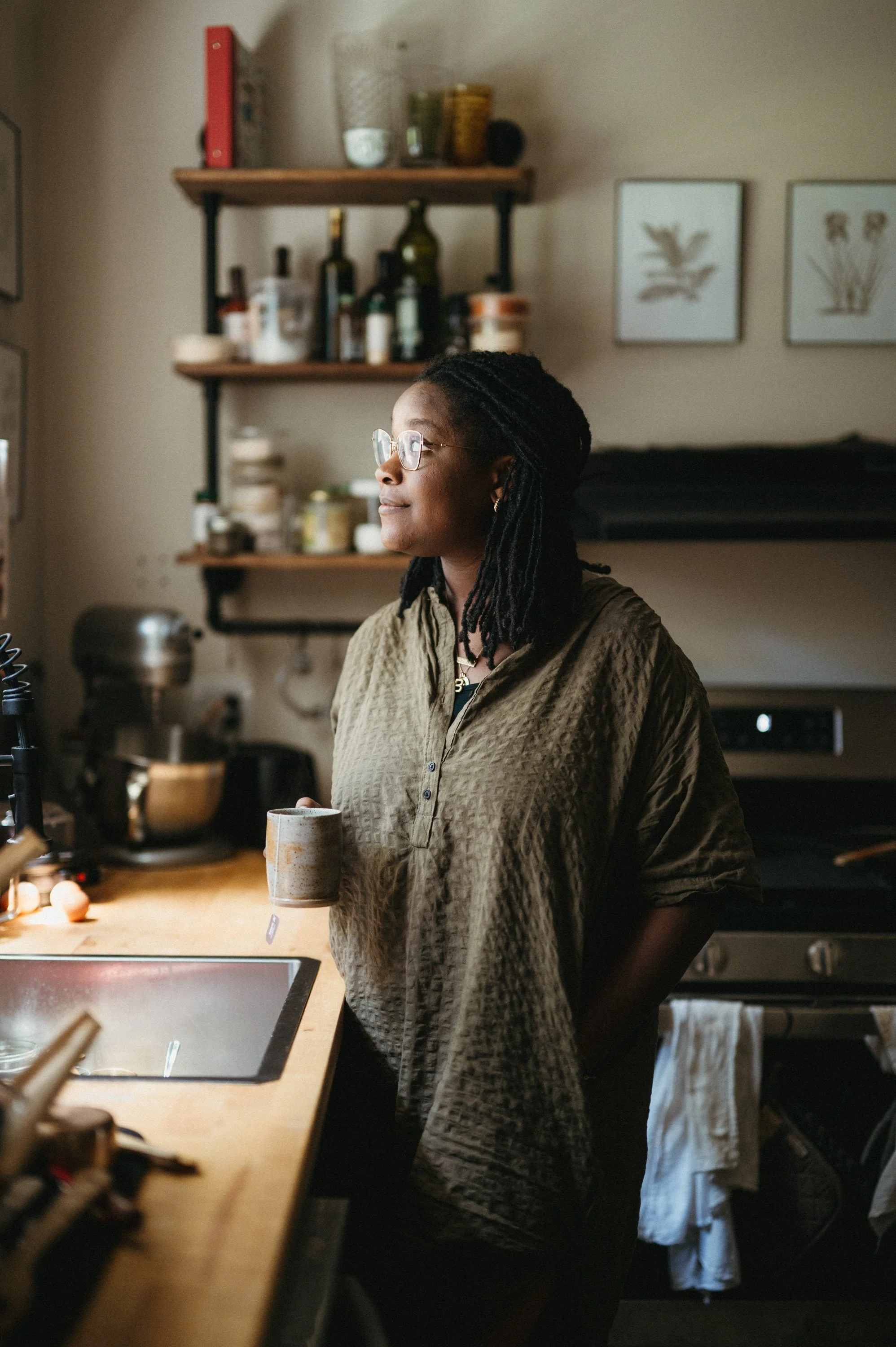 A woman standing in a kitchen, holding a coffee mug, looking out of the window, with shelves of jars and bottles behind her.