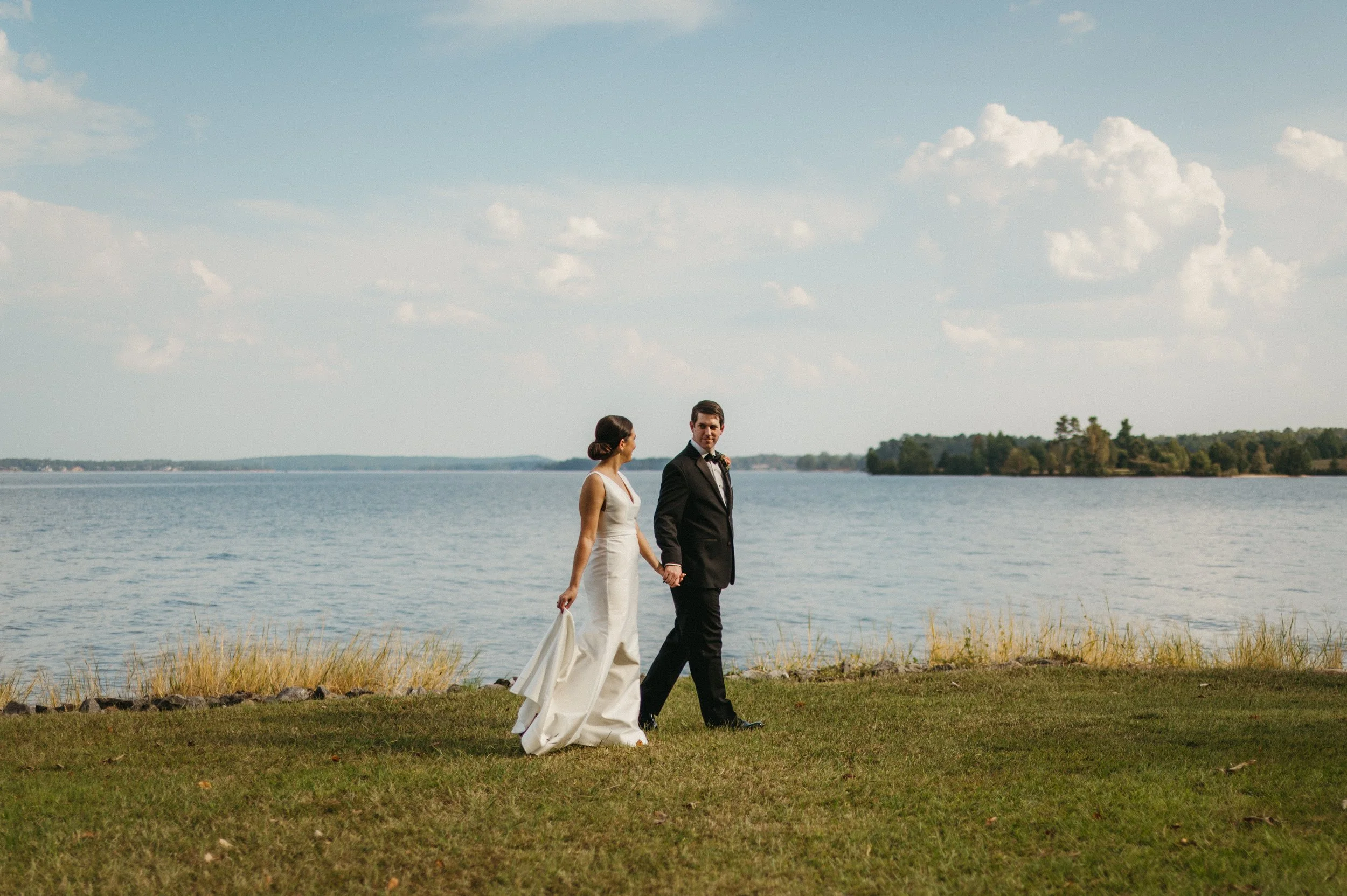 A bride and groom holding hands and walking along the grass near a lake on their wedding day, with clouds in the sky.