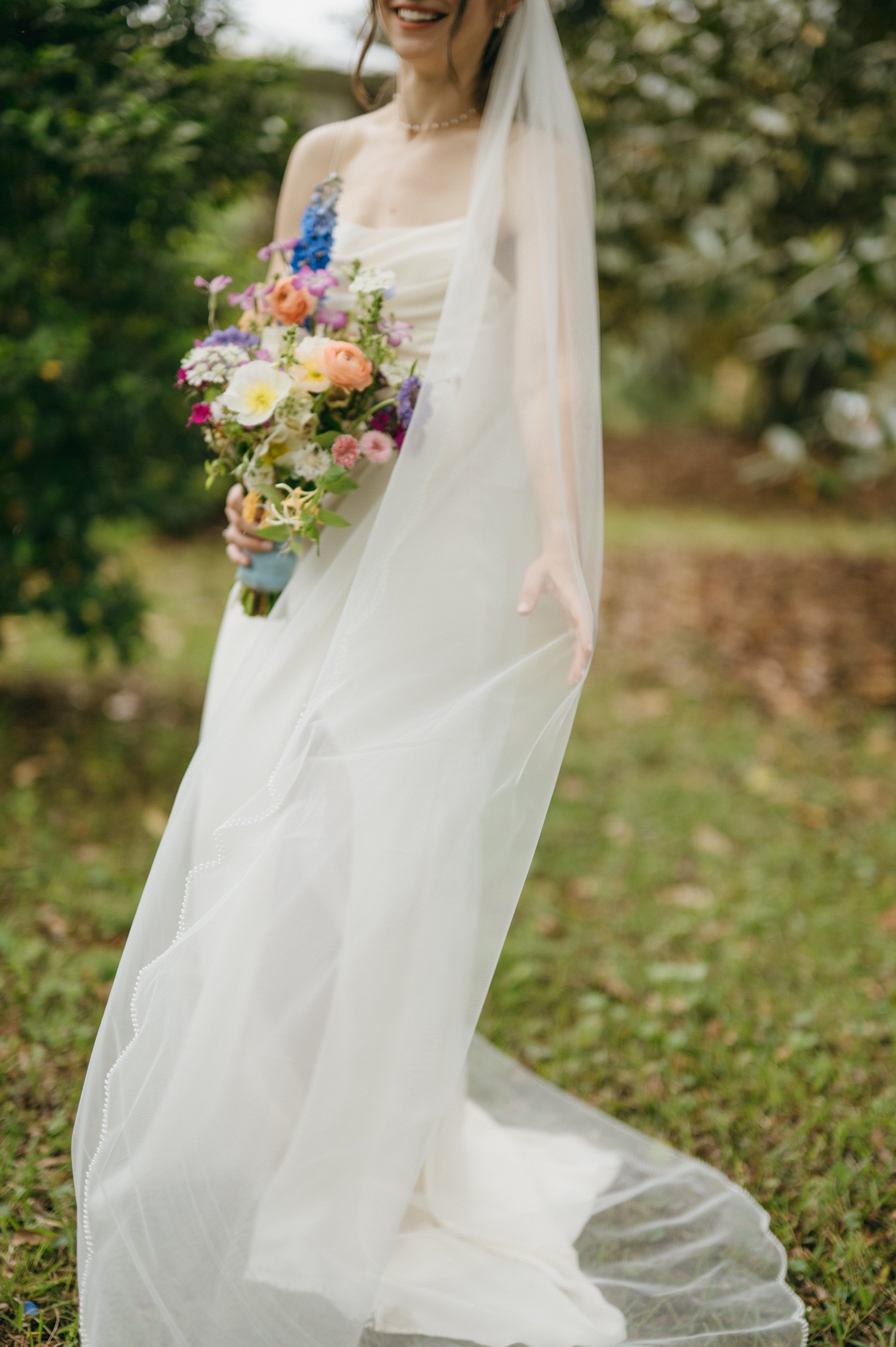 A woman in a white wedding dress holding a colorful bouquet of flowers outdoors, with a sheer veil covering part of her face and body.