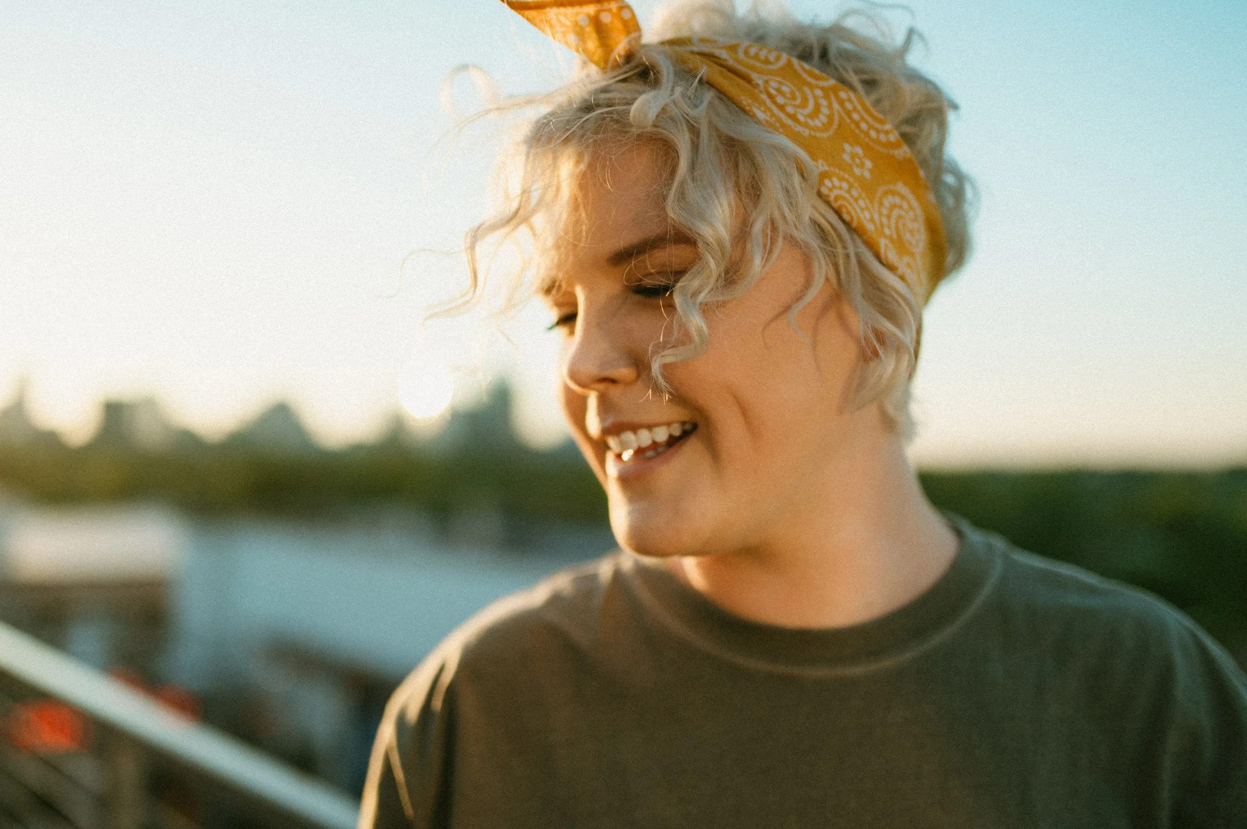 A woman with short, curly blonde hair tied with a yellow bandana, smiling outdoors during sunset.
