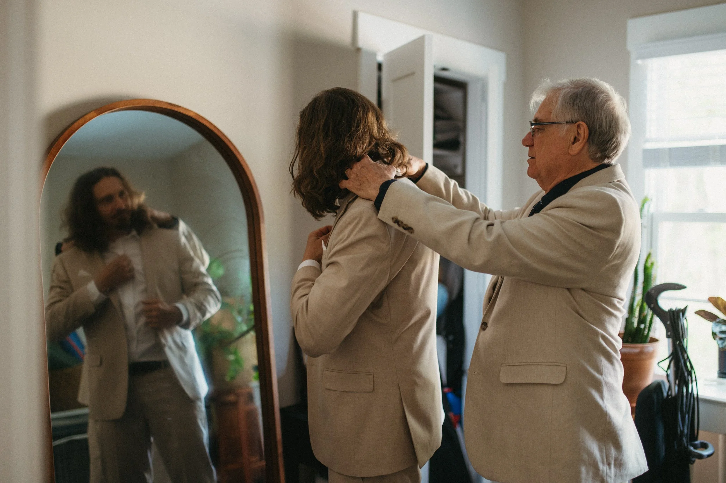 An elderly man helping a woman adjust her blazer in front of a mirror in a brightly lit room.