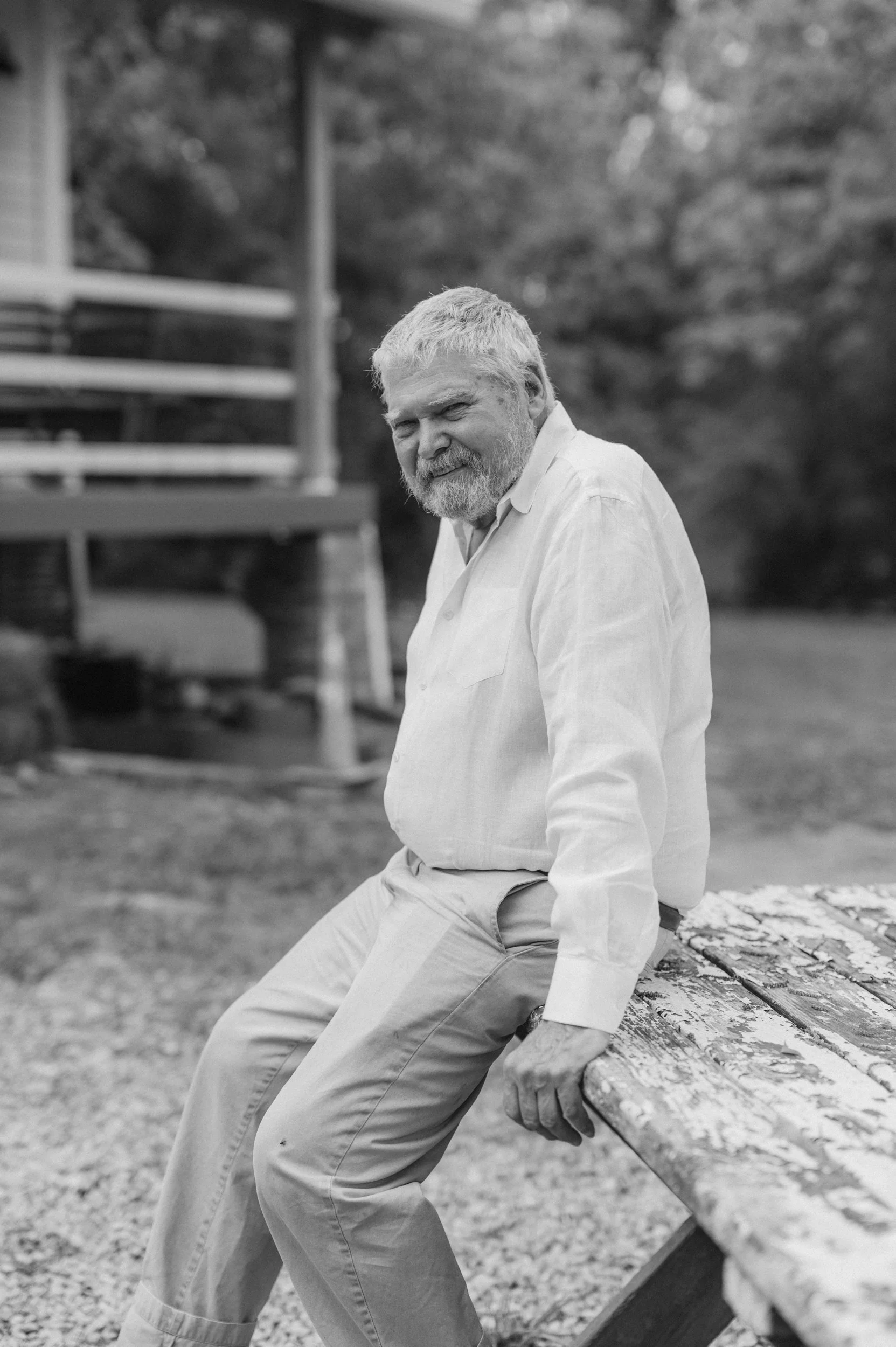 An elderly man with a beard and gray hair sitting on a weathered wooden bench outdoors, smiling and looking at the camera. He is wearing a light-colored shirt and pants, with a background of trees and a house.