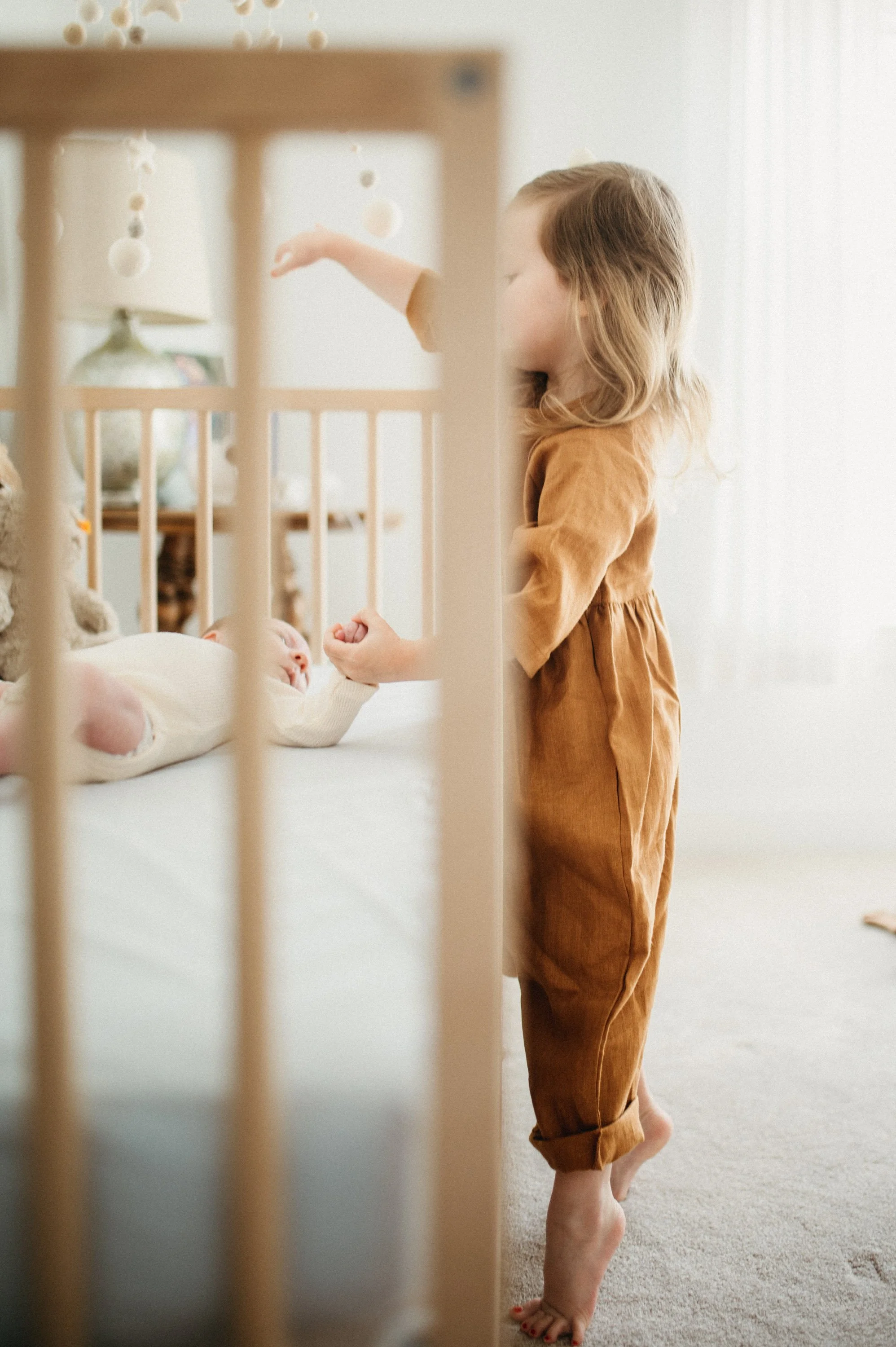 A young girl with long blonde hair standing barefoot by a crib, looking at a baby lying in the crib. The girl is wearing a brown dress or jumpsuit, and the baby is dressed in a white onesie.