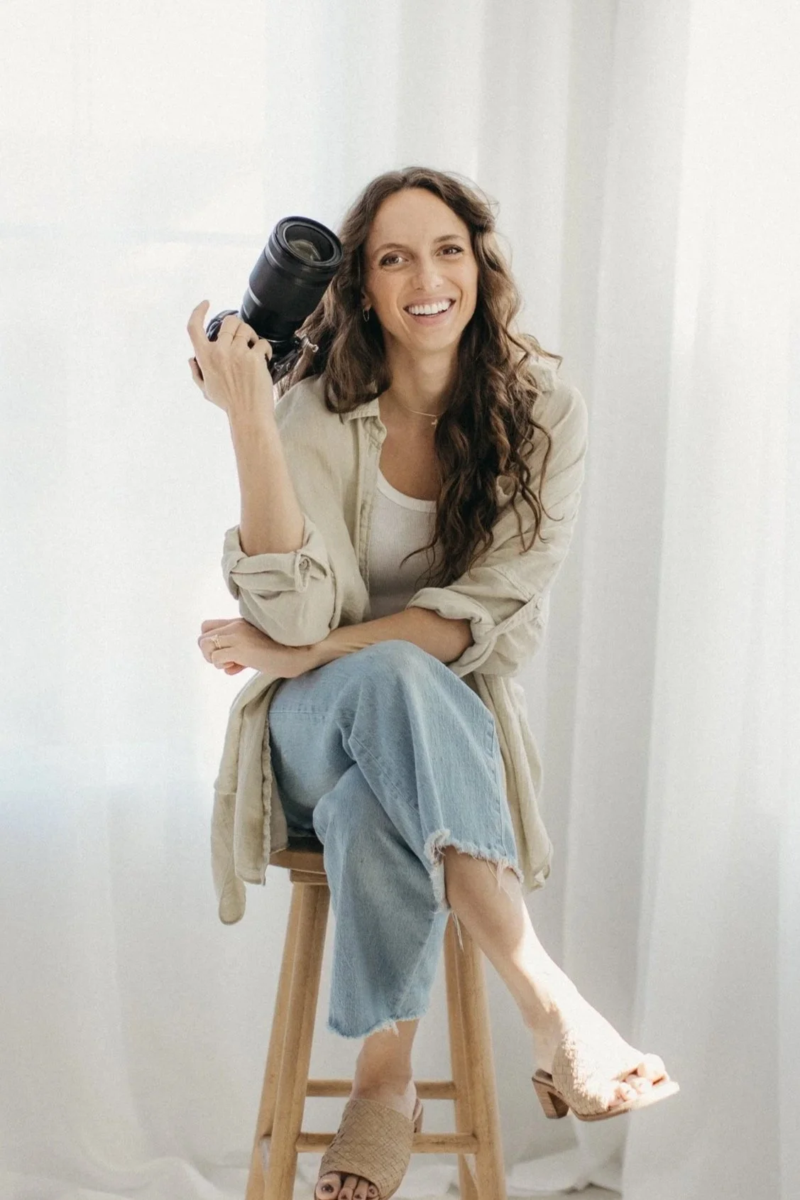 A woman with long, wavy brown hair smiling while sitting on a wooden stool, holding a camera in her right hand, wearing a white tank top, beige shirt, light blue jeans, and beige open-toed heels, with a neutral background.
