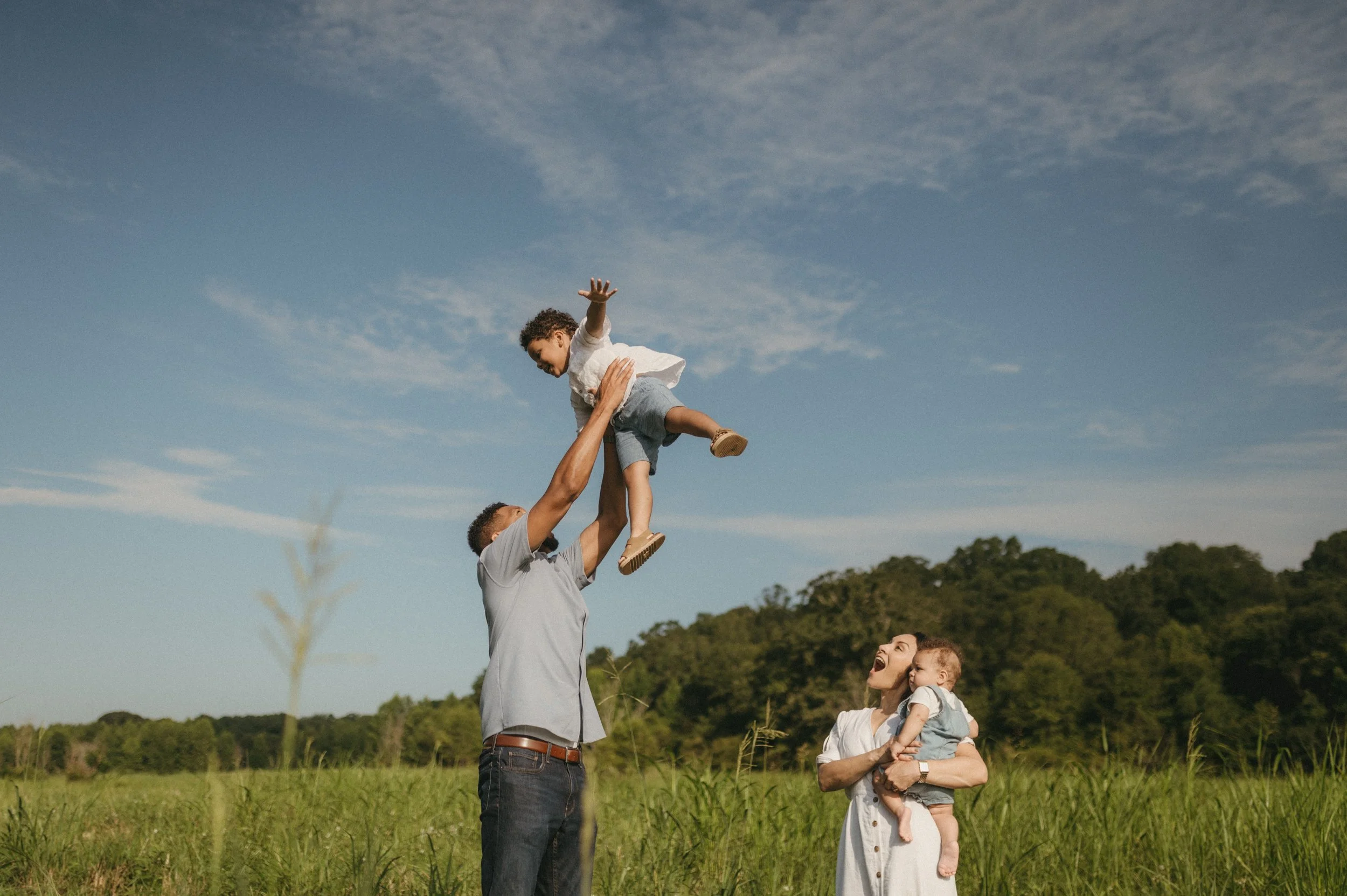 A family of four outdoors in a grassy field with trees and a blue sky, with a man lifting a young boy into the air, a woman holding a small child, and all smiling.