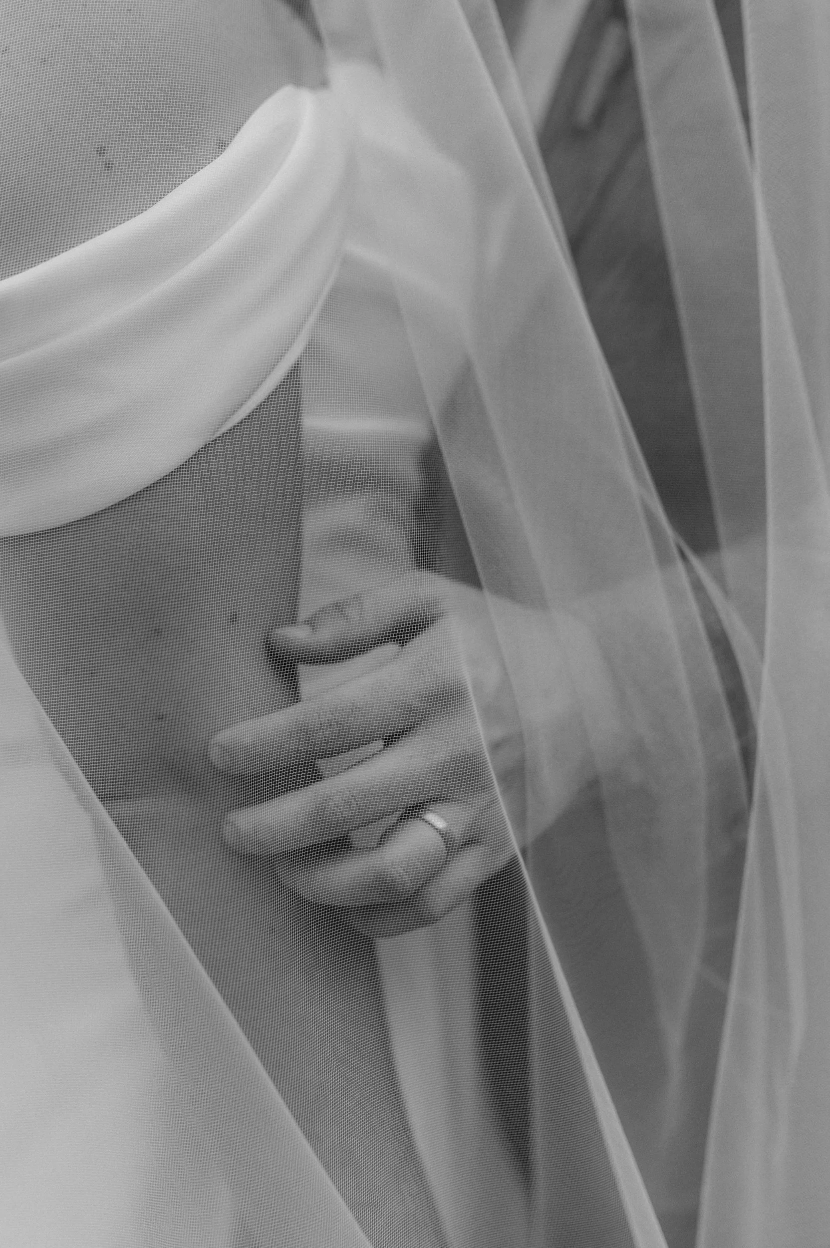 Black and white photo of a person behind a sheer veil, with only part of their face visible, and hands gently covering their mouth.