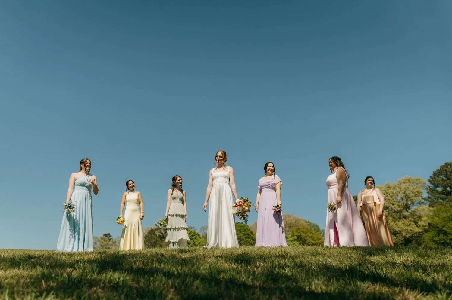 Group of young women standing on grass wearing long pastel dresses with a blue sky behind them