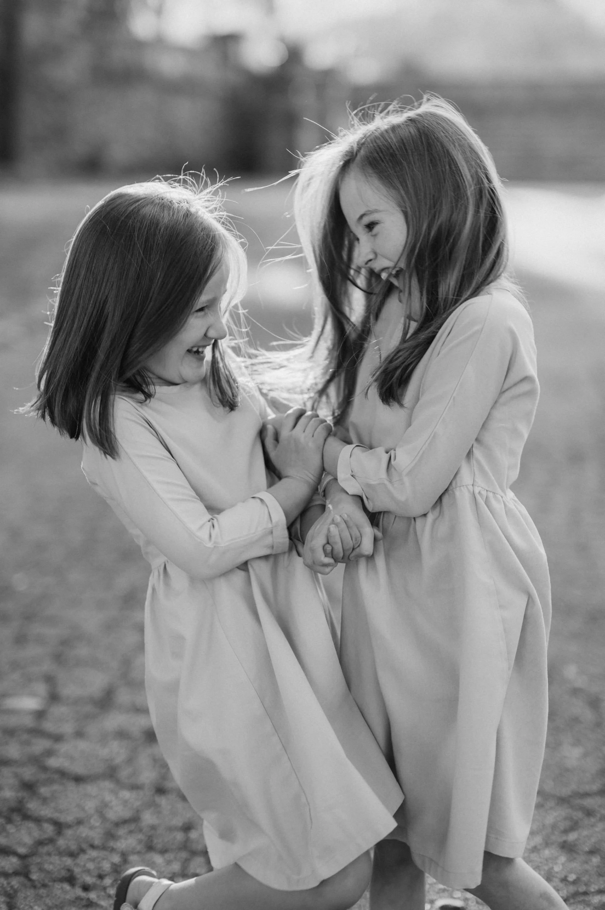 Two young girls in dresses are playing tagether outside, holding hands and laughing.