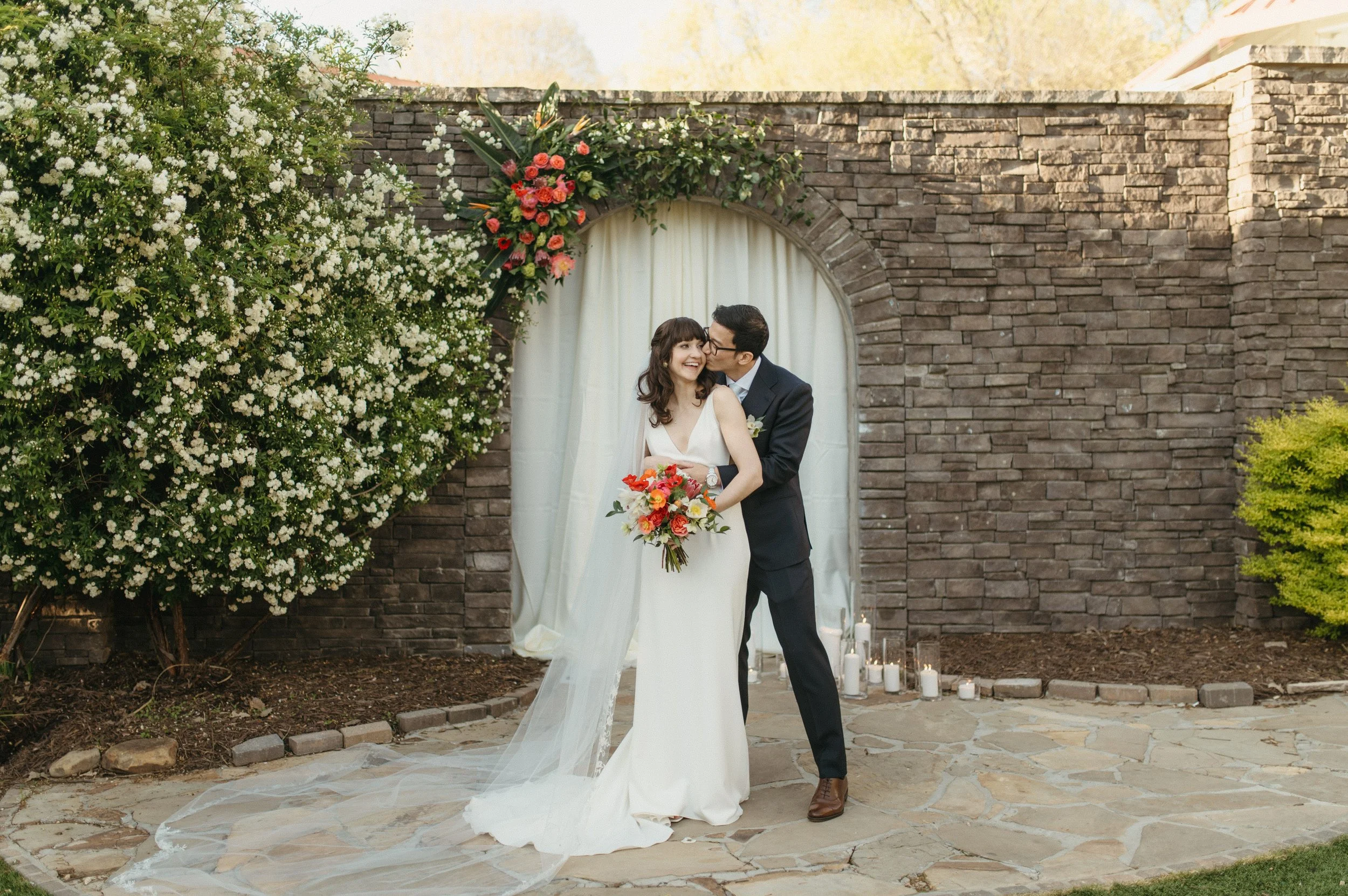 Bride and from stand close and smile in front of a stone wall covered in flowers